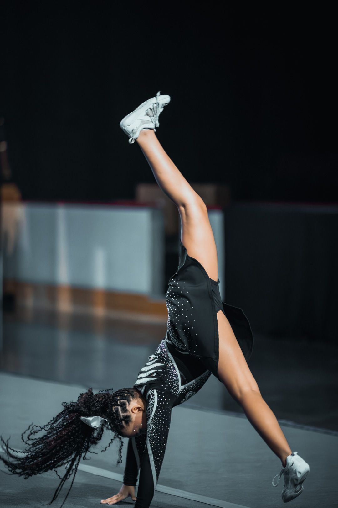 Young gymnast in black leotard performs a side aerial split on stage, one leg raised high, wearing white sneakers.
