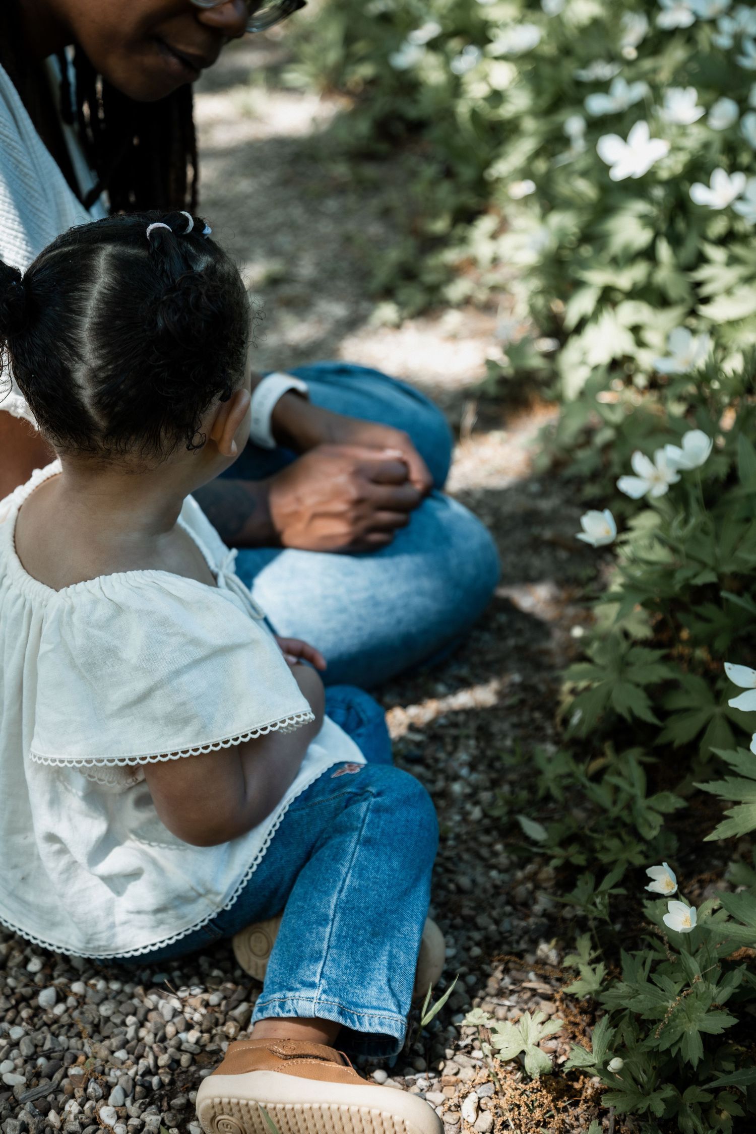 A parent and young child sit together on a gravel path, exploring white flowers in a lush garden.