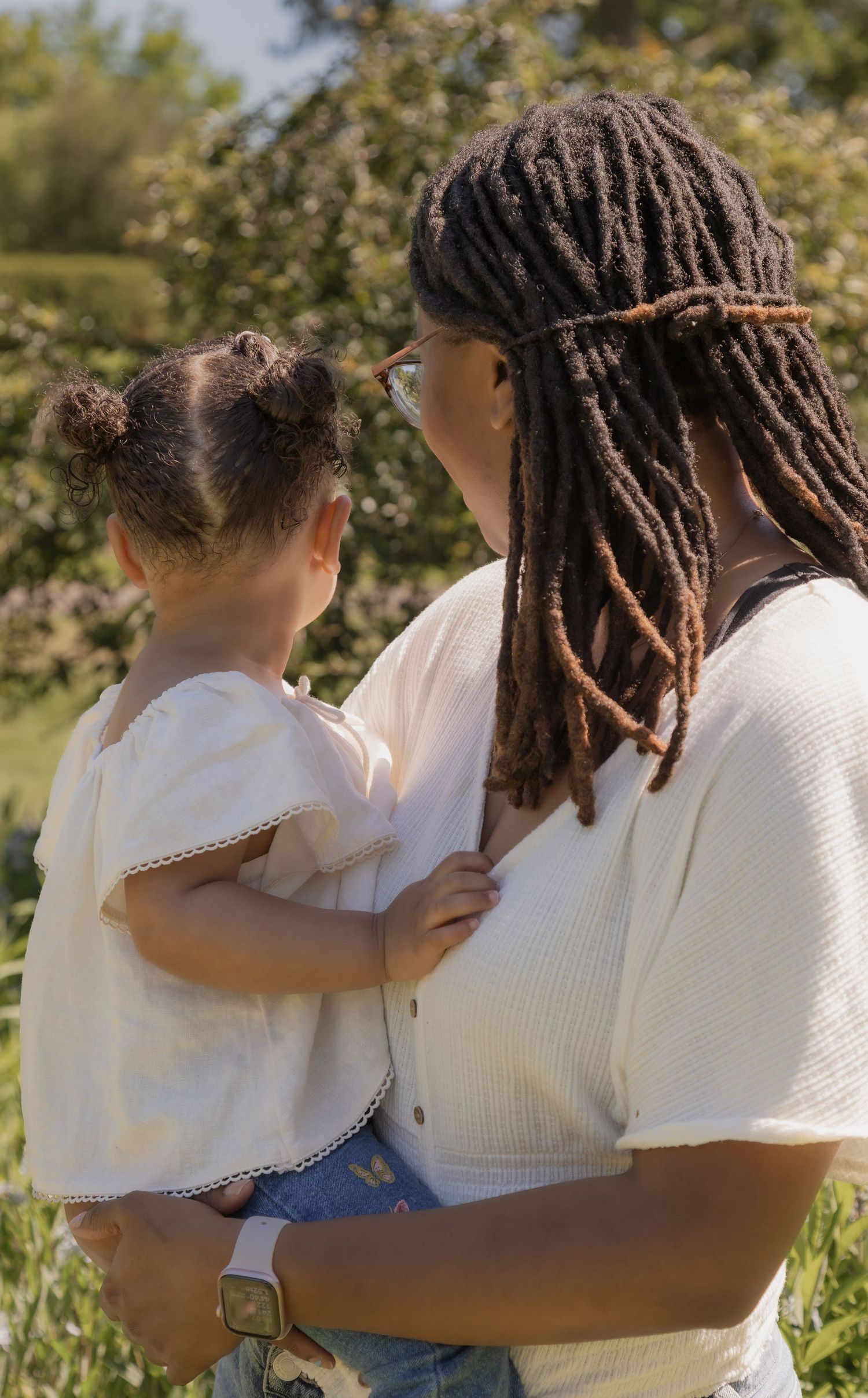 A mother with locs holds her young daughter in a sunny garden, both wearing white tops.