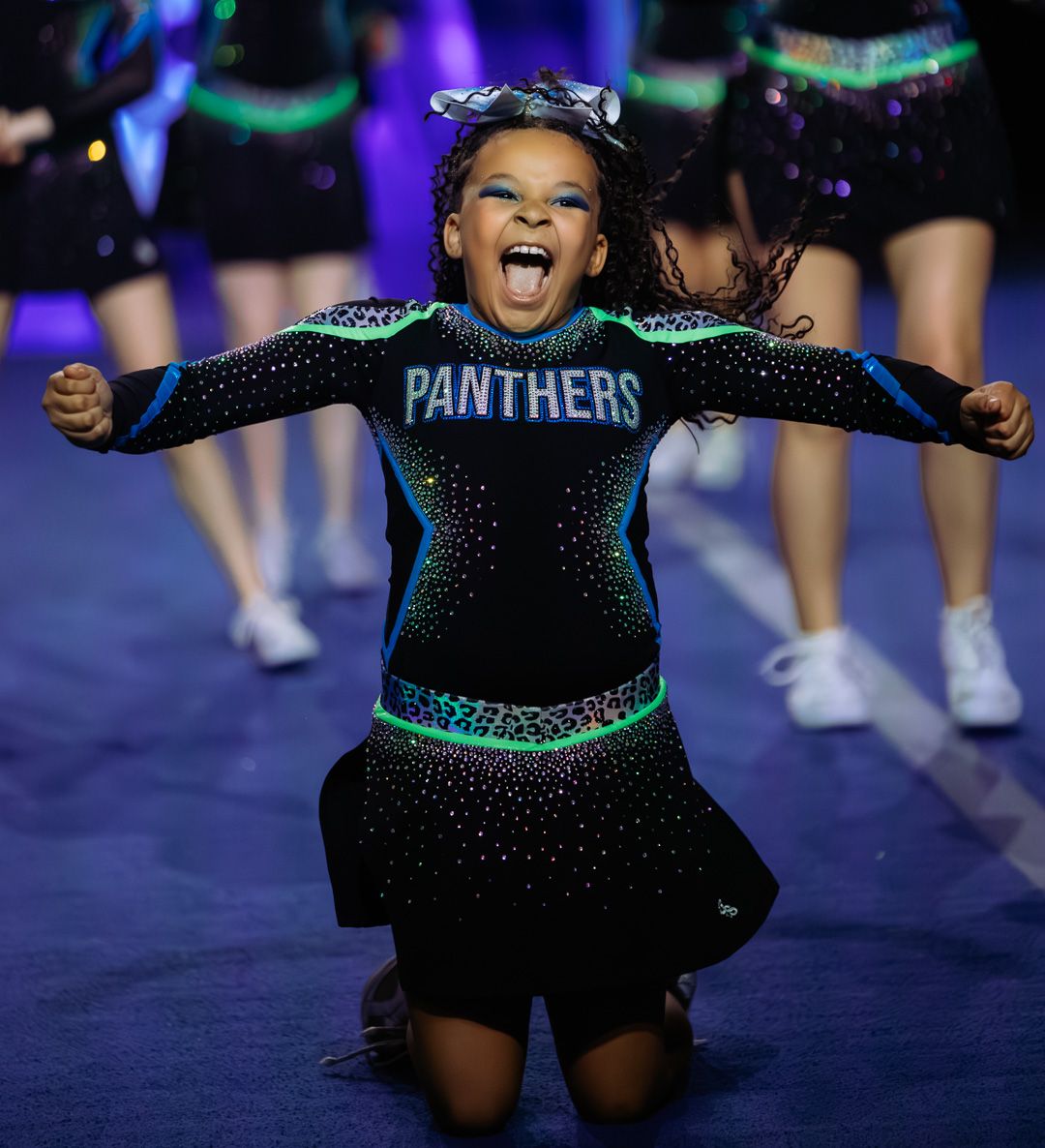 Young cheerleader in black Panthers uniform performs energetically on stage, arms raised with excitement.