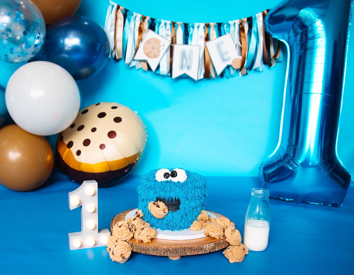 Cookie Monster themed first birthday party setup with blue cake, cookies, balloon number 1, and ONE banner.