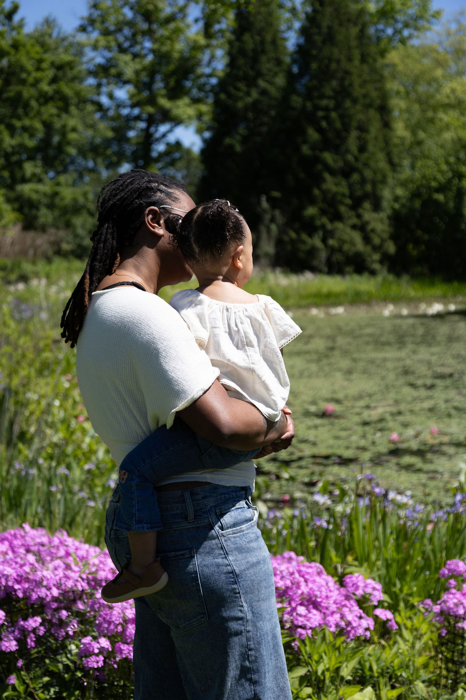 A mother with dreadlocks holds her young child while standing near a lily pond surrounded by purple flowers.