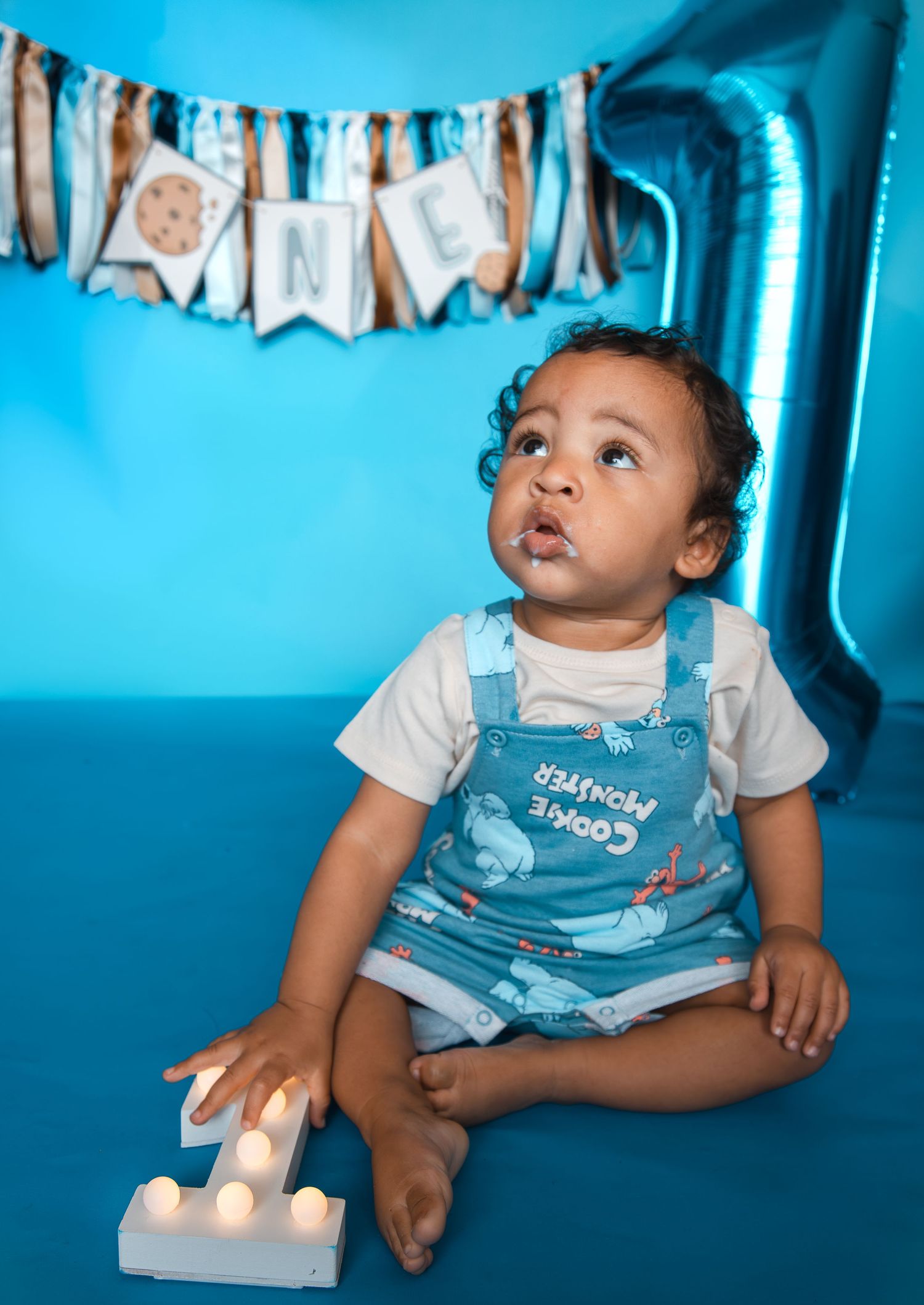 Baby in Cookie Monster overalls sits on blue floor celebrating first birthday with ONE banner and balloon.