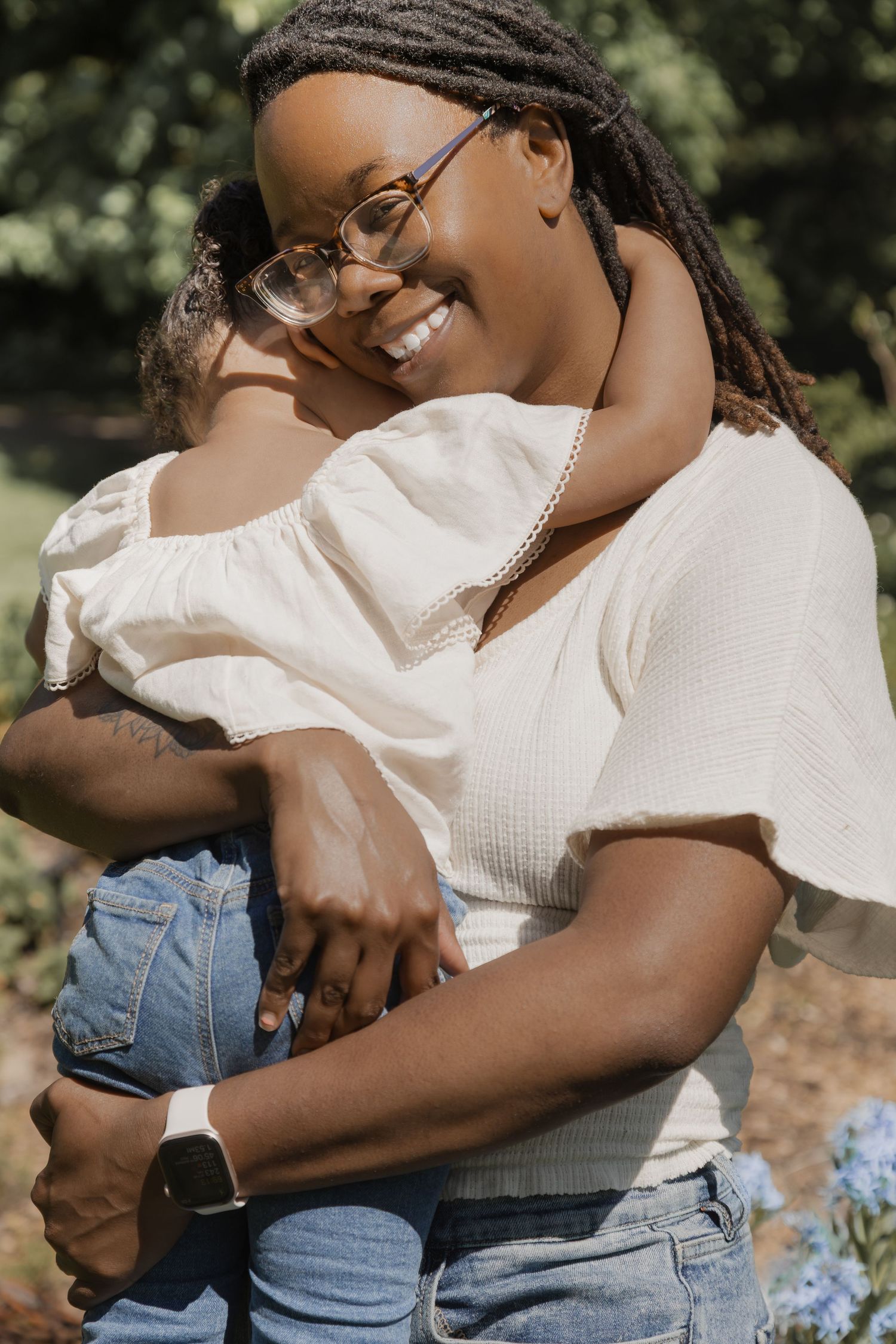 Smiling Black mother with dreadlocks and glasses hugging her young daughter outdoors in a garden.