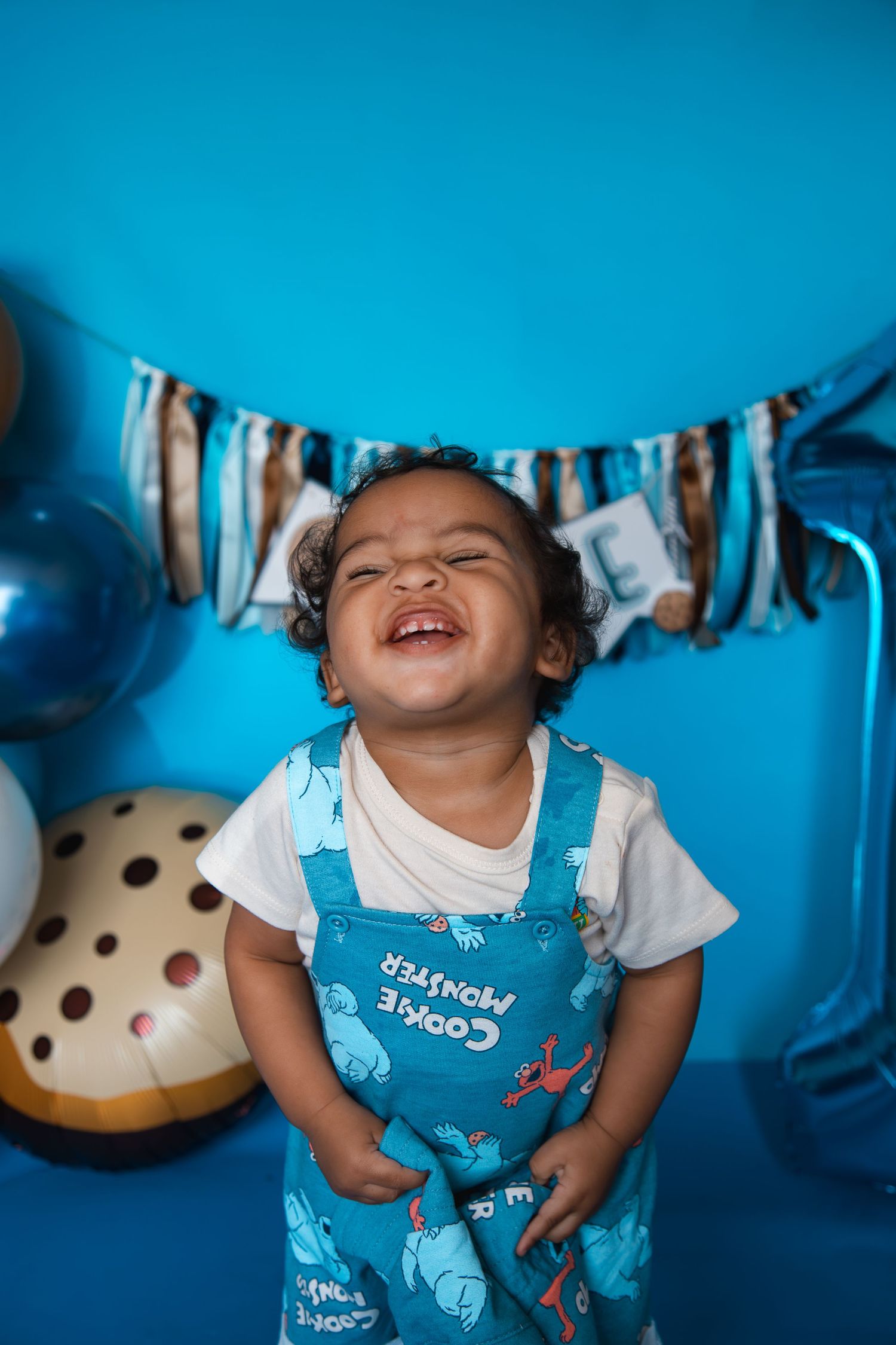 Joyful toddler in Cookie Monster overalls laughing at a blue-themed first birthday party with balloons and bunting.