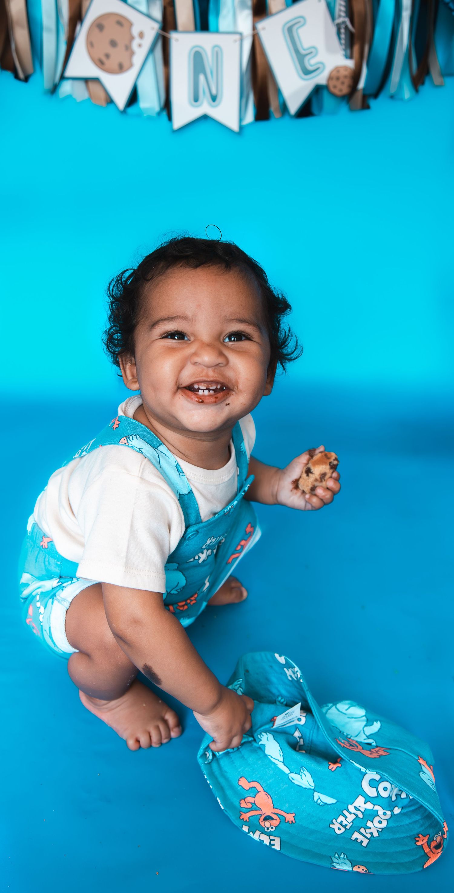 Smiling baby boy in teal Cookie Monster overalls celebrating his first birthday on a blue background.