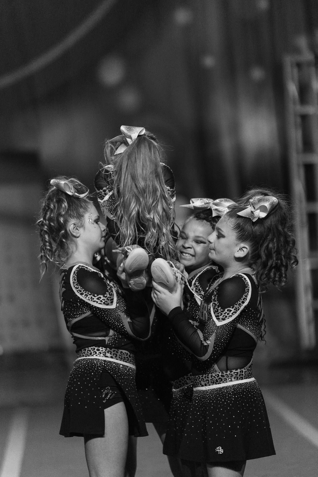 Young cheerleaders in sparkly uniforms with hair bows celebrate together at a cheerleading competition in black and white photo.