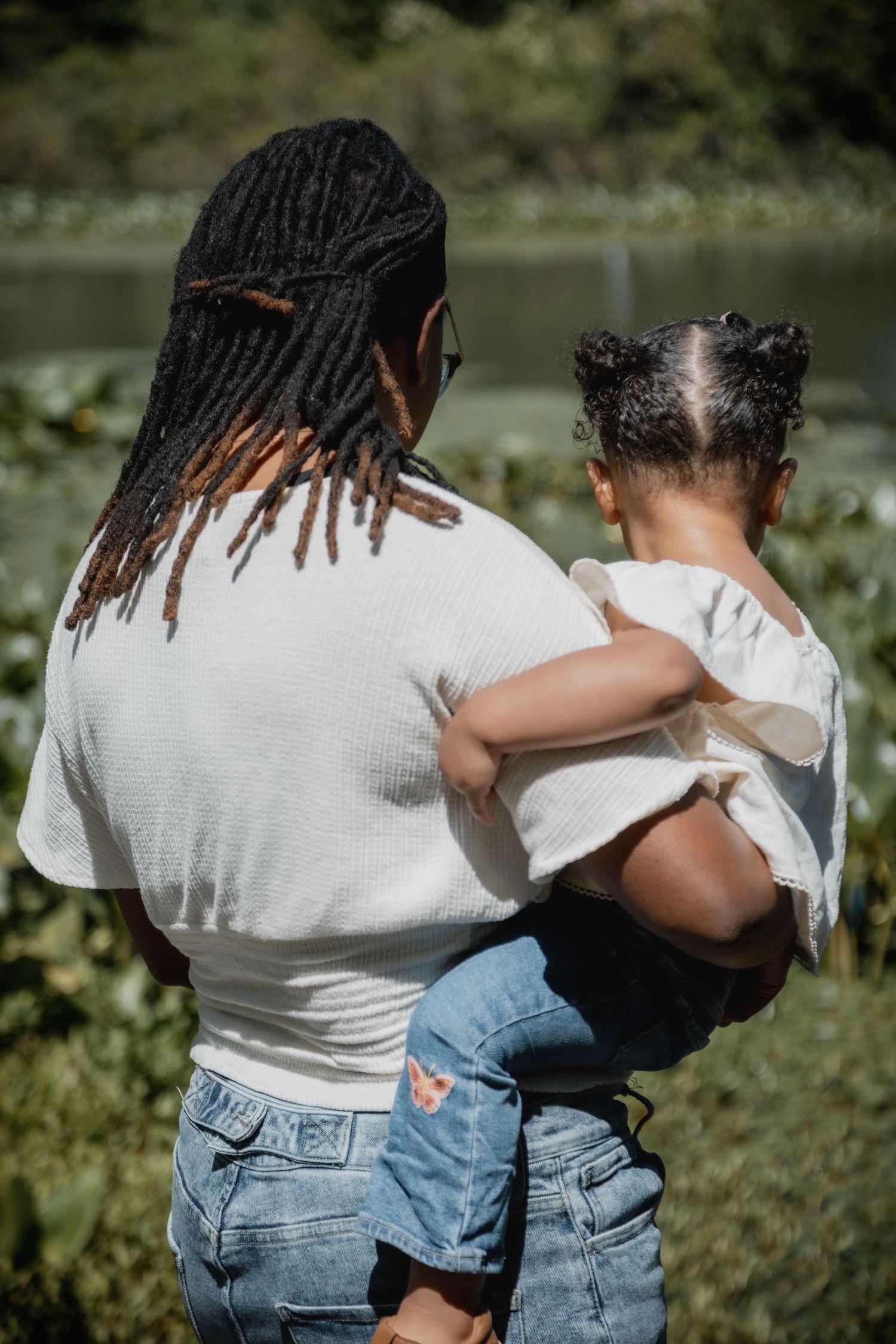 A mother with dreadlocks holds her young child while looking out over a serene pond surrounded by greenery.