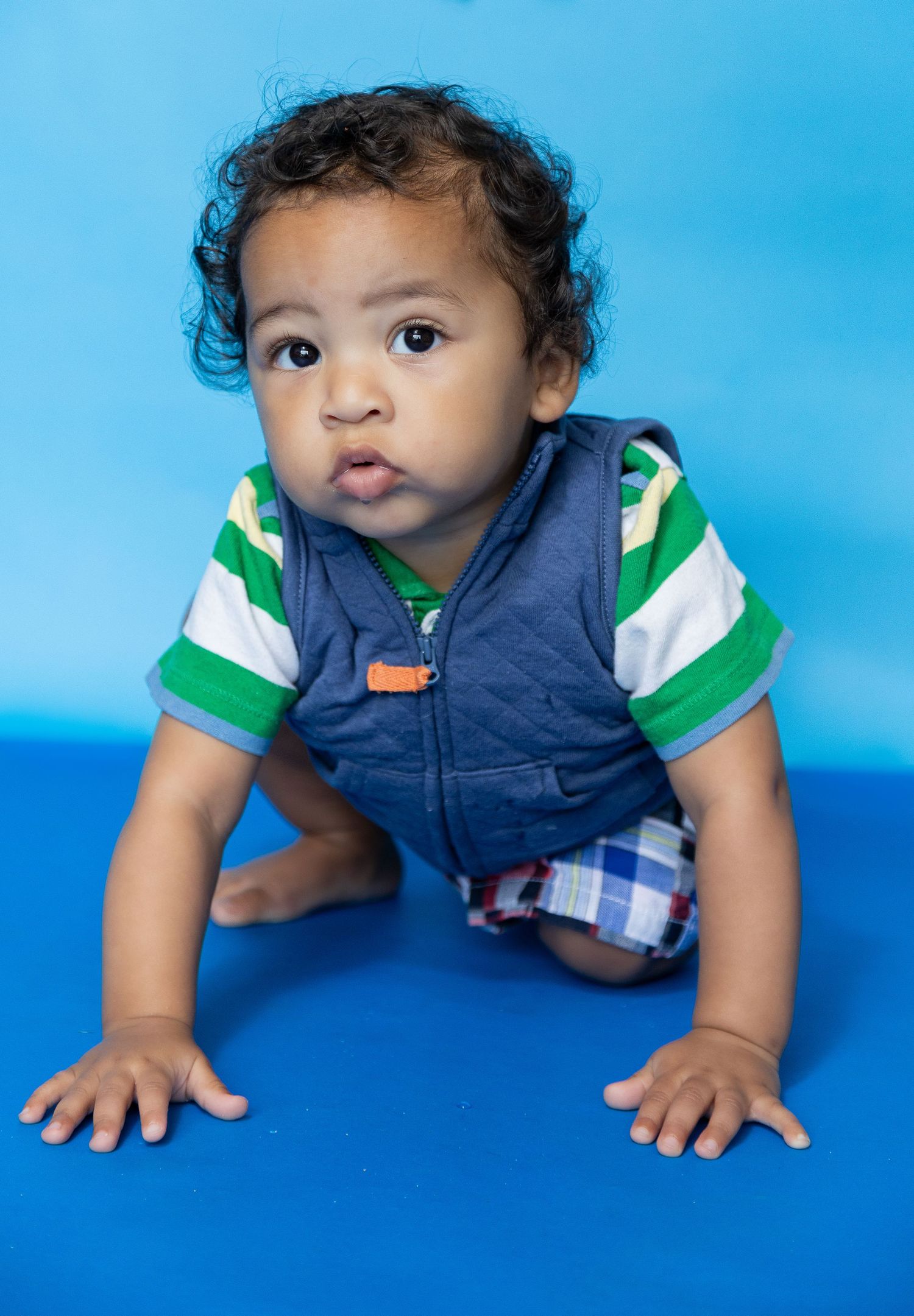 Adorable baby boy in navy vest and striped shirt crawling on blue floor, looking at camera with curly hair.