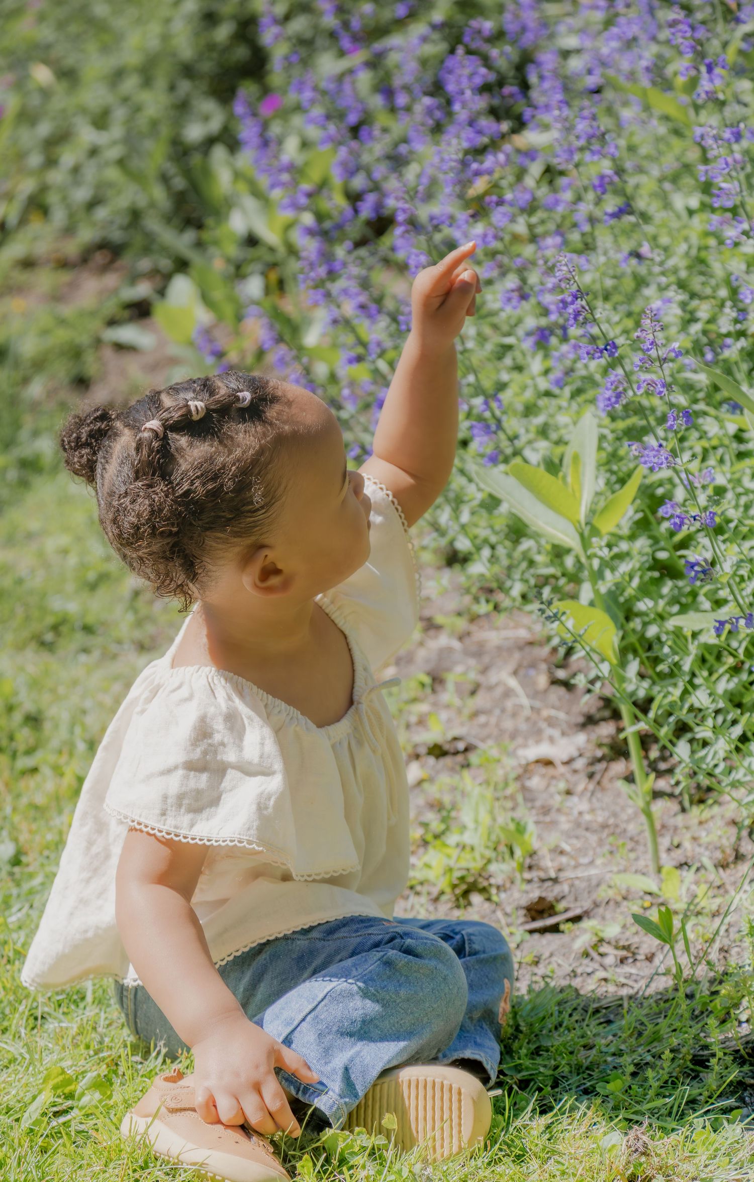 Toddler girl in white top and jeans sitting on grass reaching for purple wildflowers in a sunny garden.