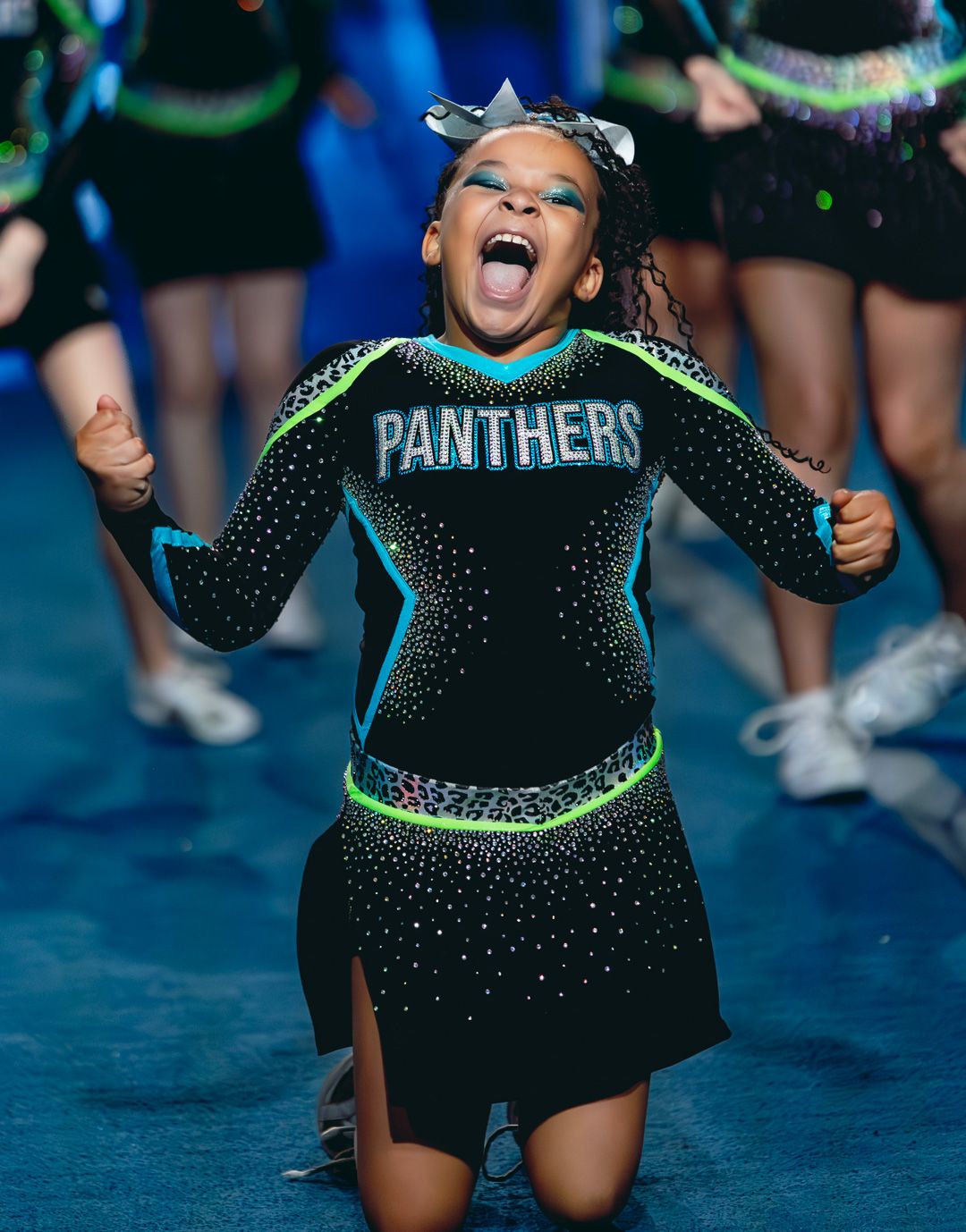 Young cheerleader in black Panthers uniform with neon trim, cheering enthusiastically on a blue competition floor.