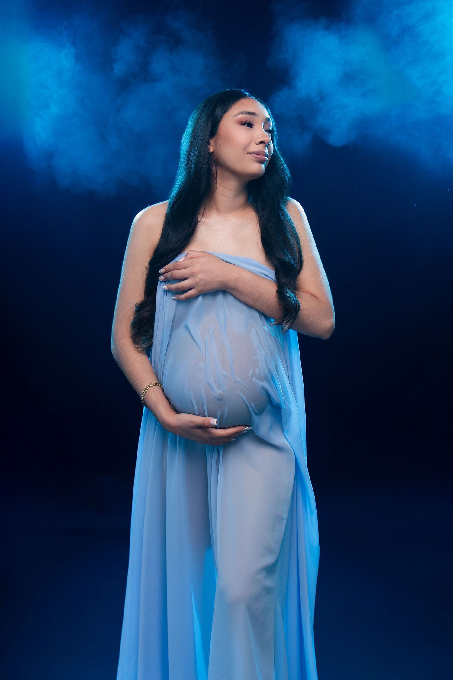 Pregnant woman in blue sheer fabric poses gracefully against a dark blue smoky background, cradling her baby bump.