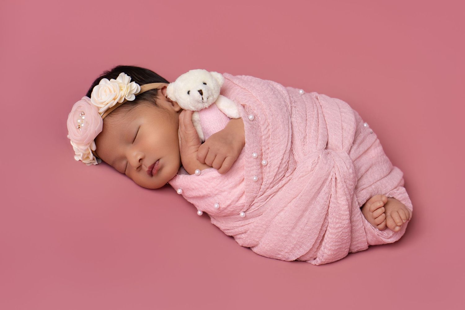 Sleeping newborn baby girl swaddled in pink wrap with floral headband holding a tiny white teddy bear.