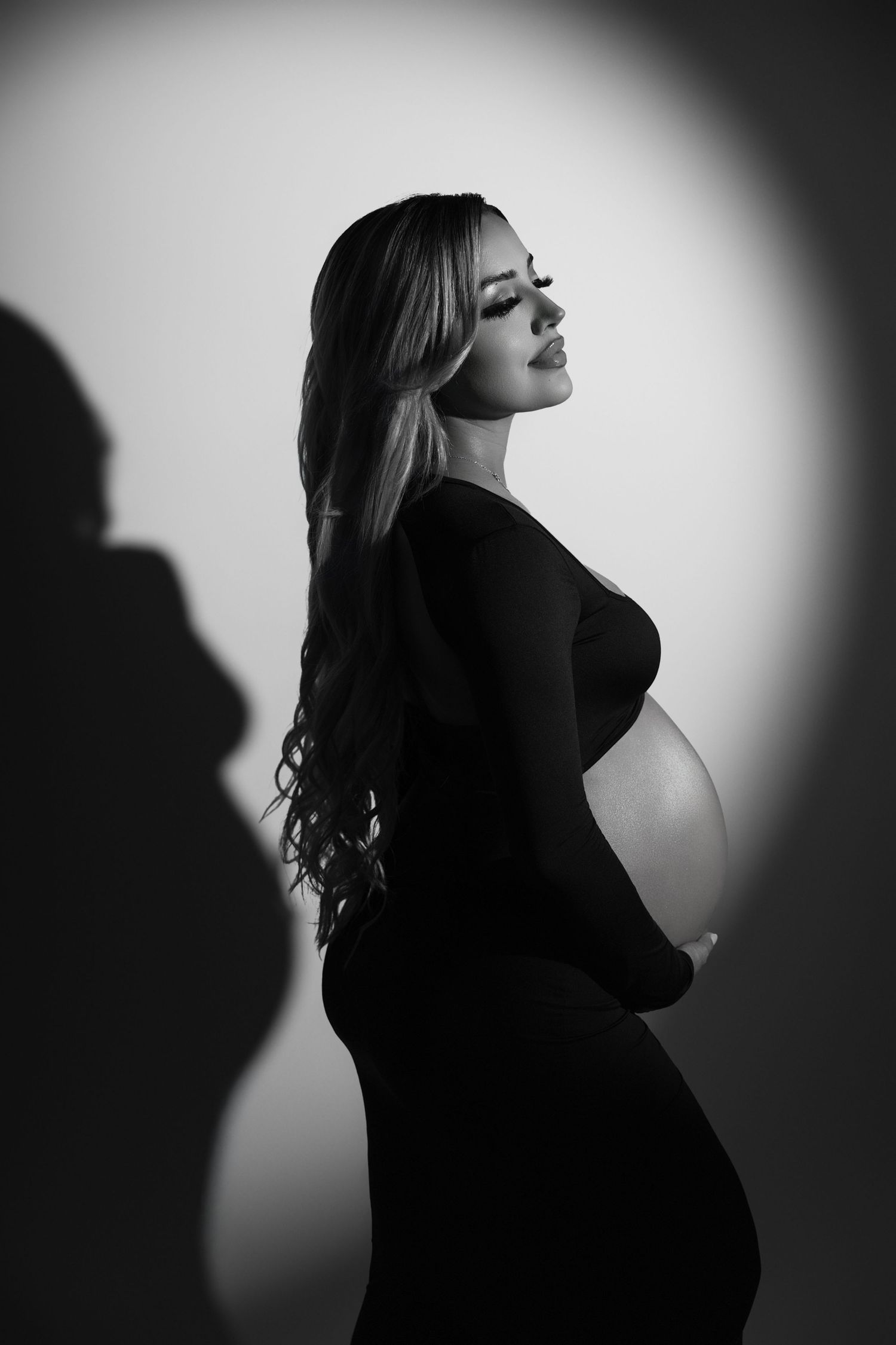 Black and white maternity photo of a pregnant woman in a black dress cradling her baby bump in dramatic studio lighting.