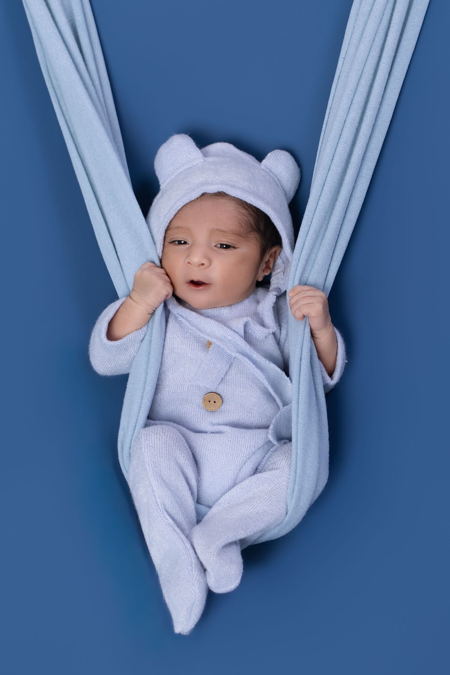 Newborn baby in light blue bear hat and onesie resting in blue fabric sling against blue background.