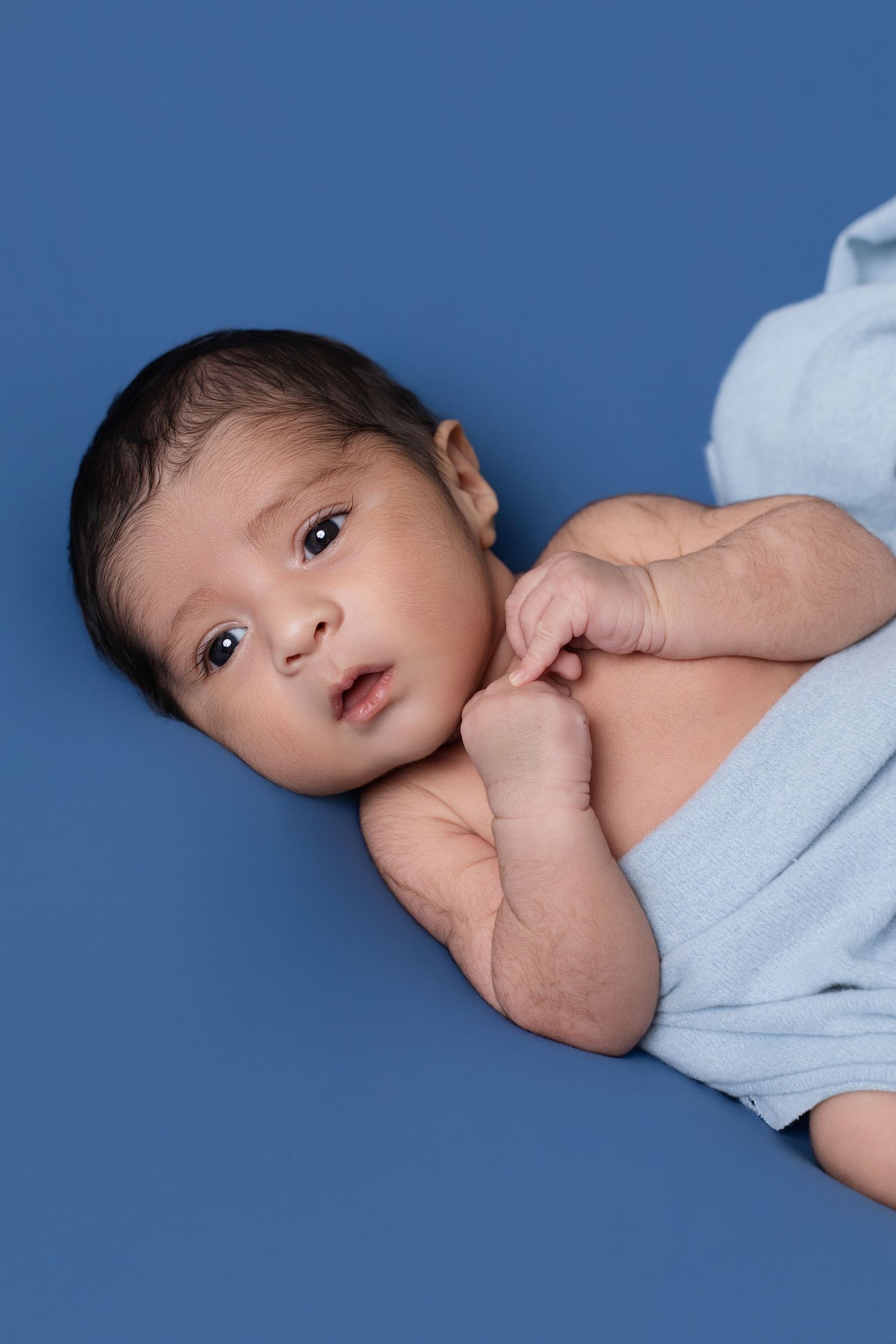 A newborn baby with dark hair lies on a blue blanket, gazing upward with tiny fists tucked under chin.
