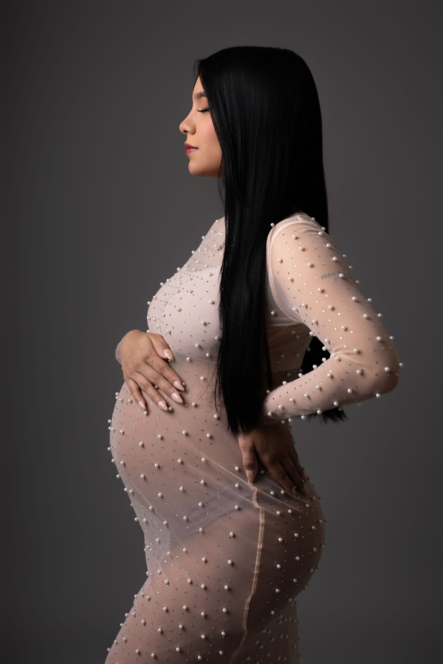 Side profile of a pregnant figure in a sheer beaded dress with long straight black hair against a dark background.