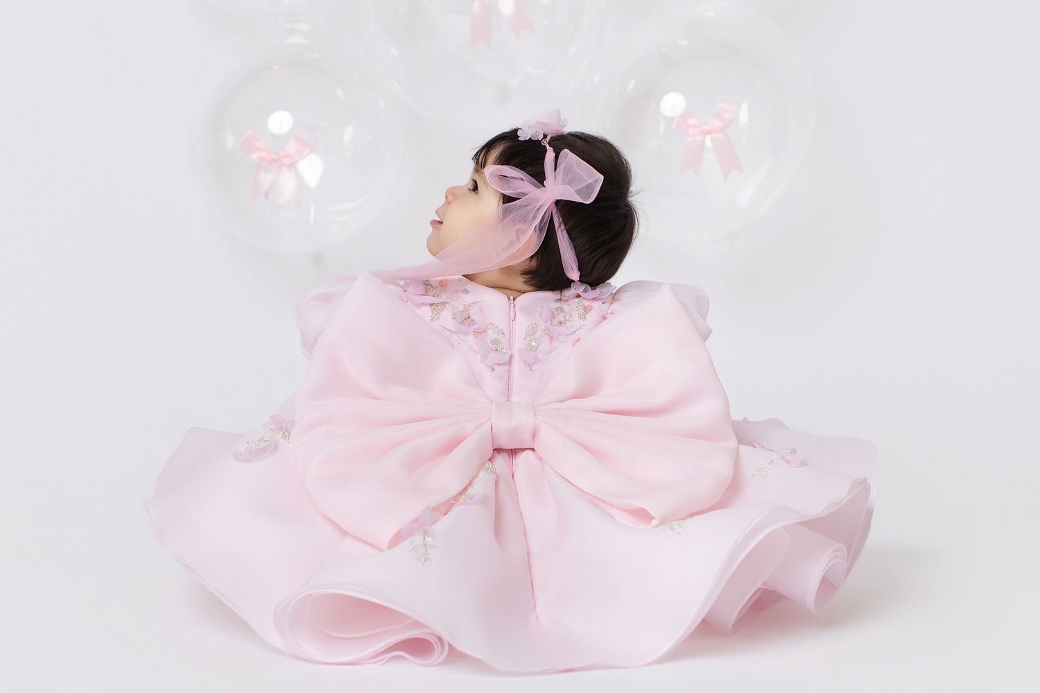 Baby girl in pink ruffle dress with large bow, wearing pink bow headband, surrounded by clear balloons.