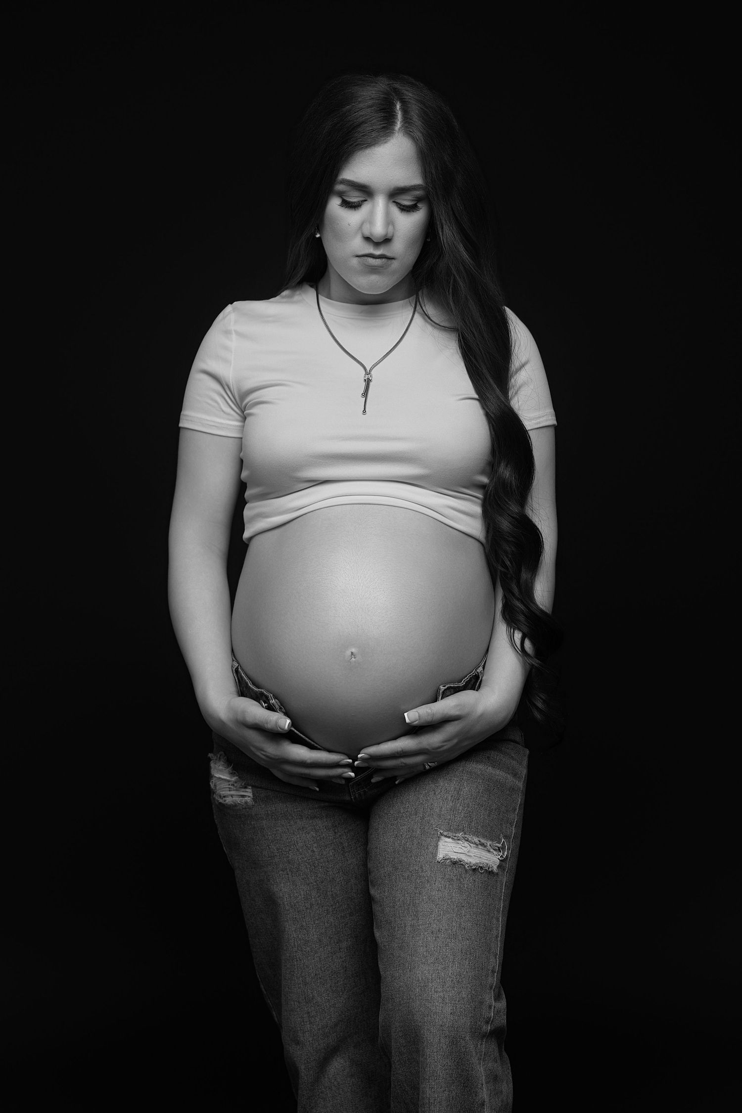 Black and white portrait of a pregnant woman cradling her bare belly, wearing a crop top and ripped jeans.