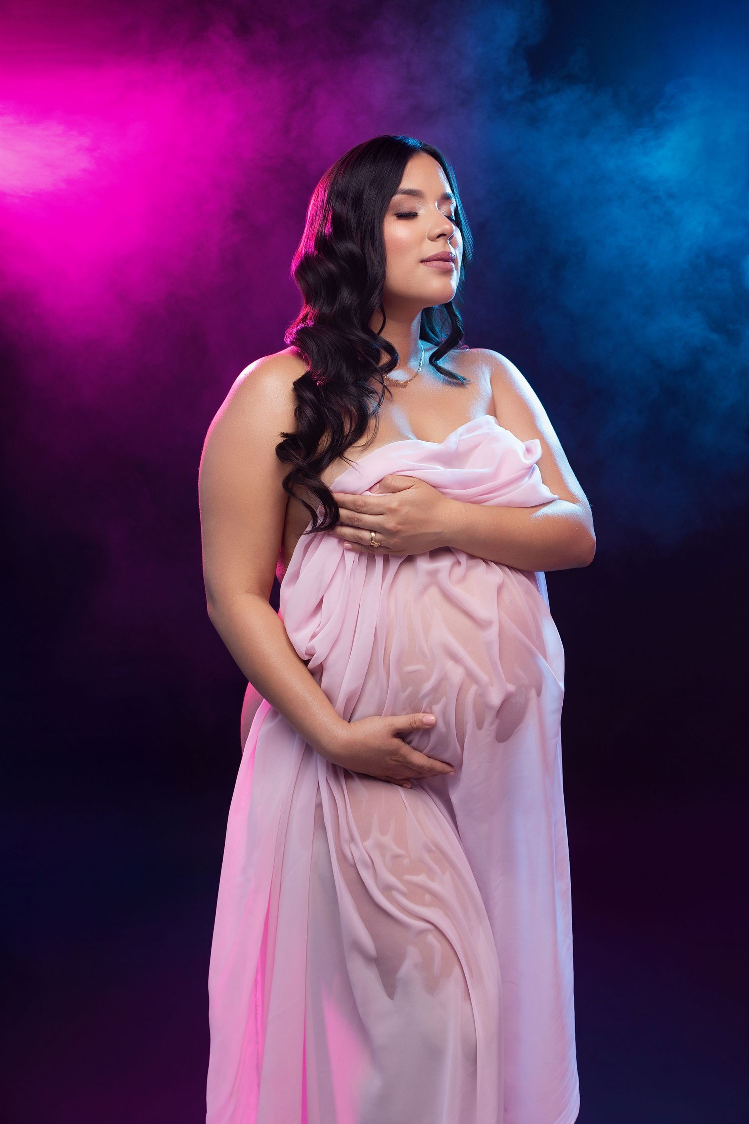 Pregnant woman in pink draped fabric poses gracefully against dramatic pink and blue smoky dark background.