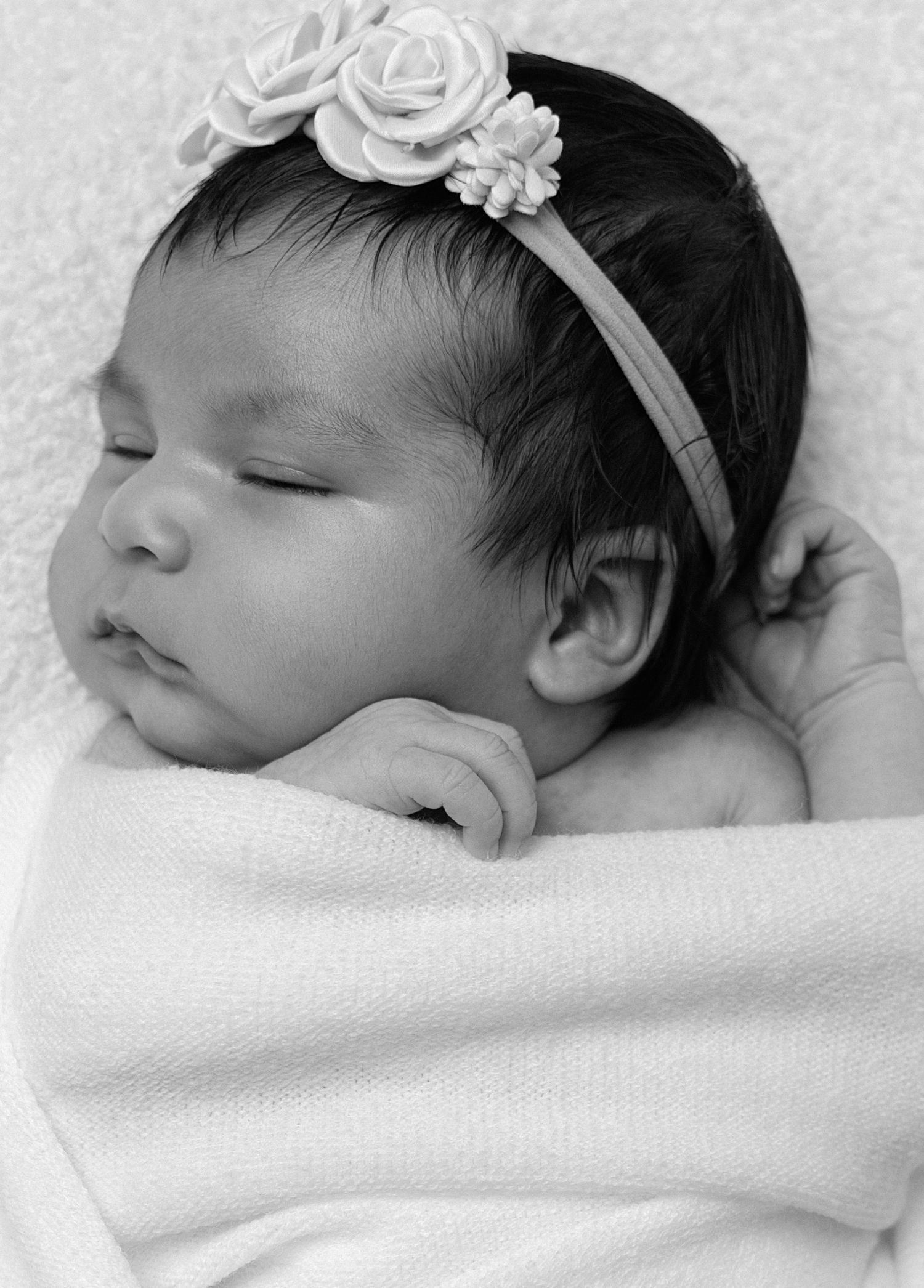 Black and white newborn baby girl sleeping peacefully, wrapped in white blanket, wearing floral headband.