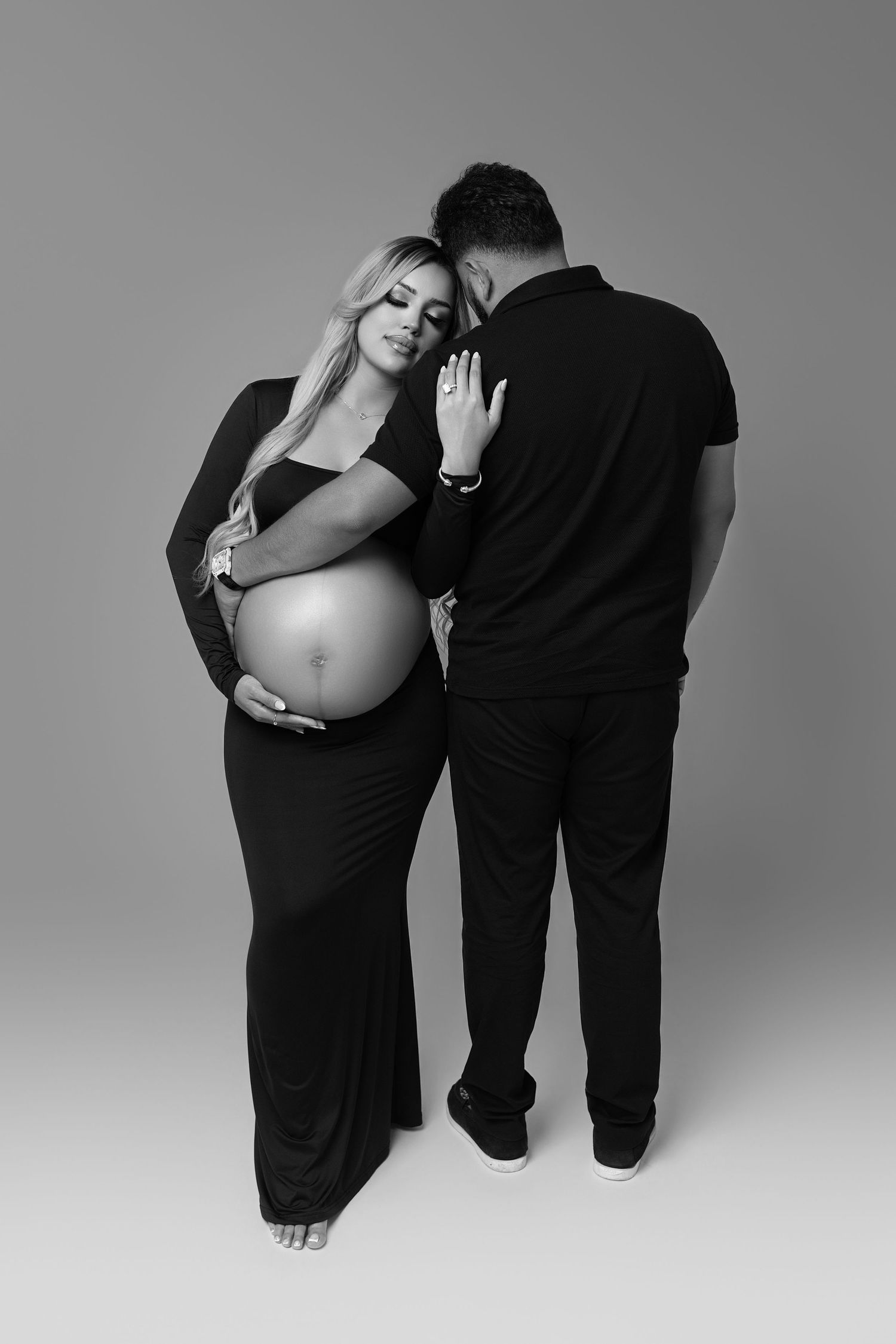 Black and white maternity photo of pregnant woman in black dress with partner in studio setting.
