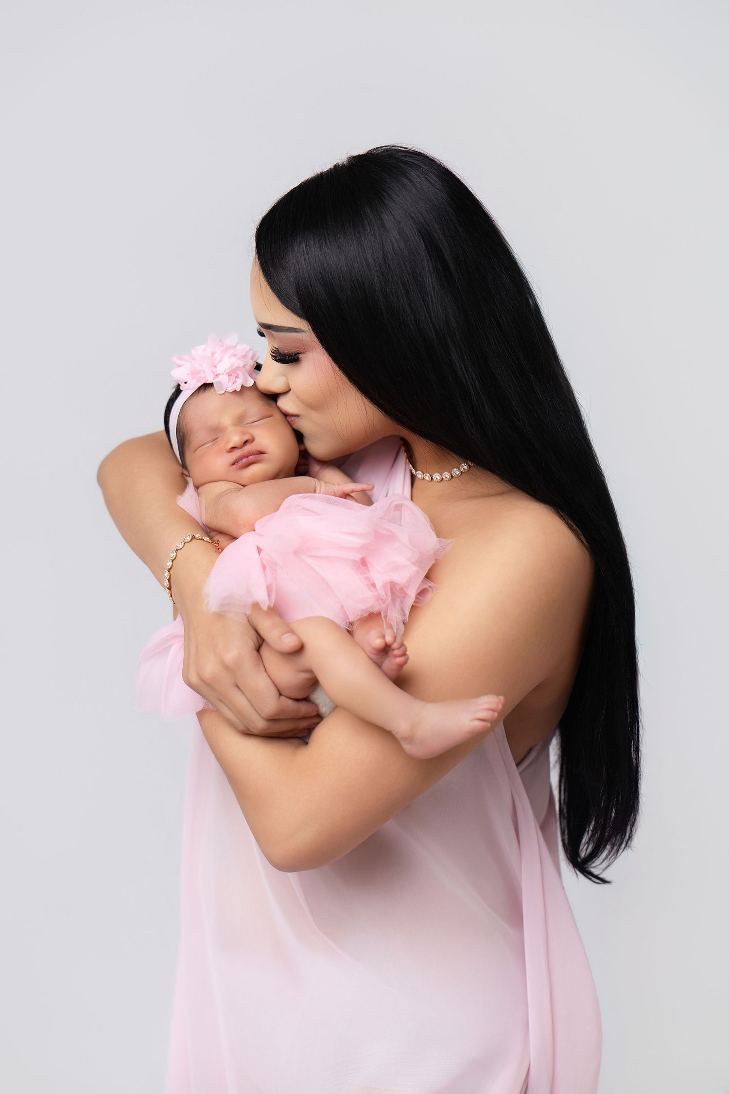A mother in pink gown kisses her newborn baby girl wrapped in pink tulle with a floral headband.