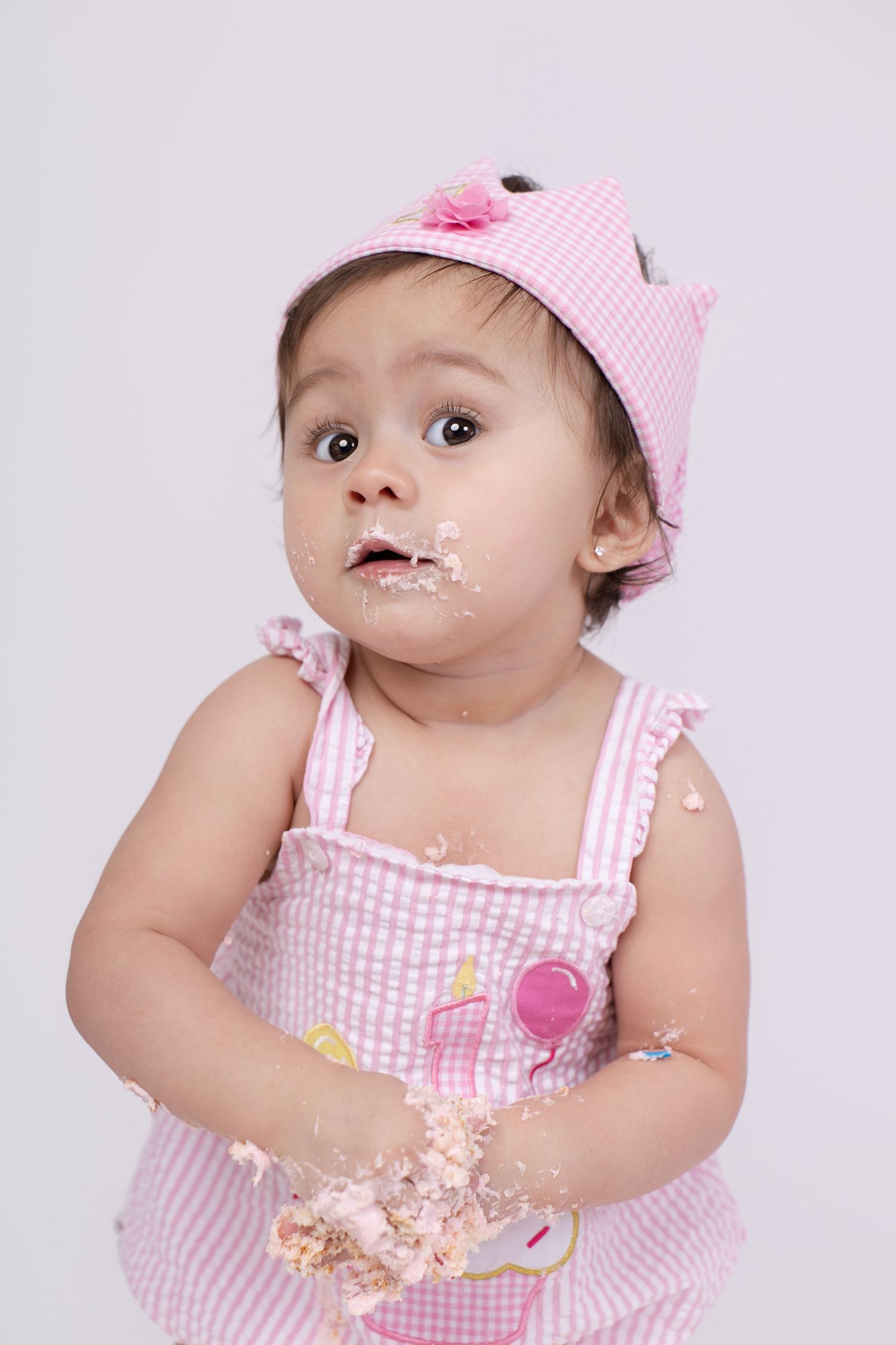 Cute baby girl in pink crown and dress with cake smash on her face and hands during first birthday.