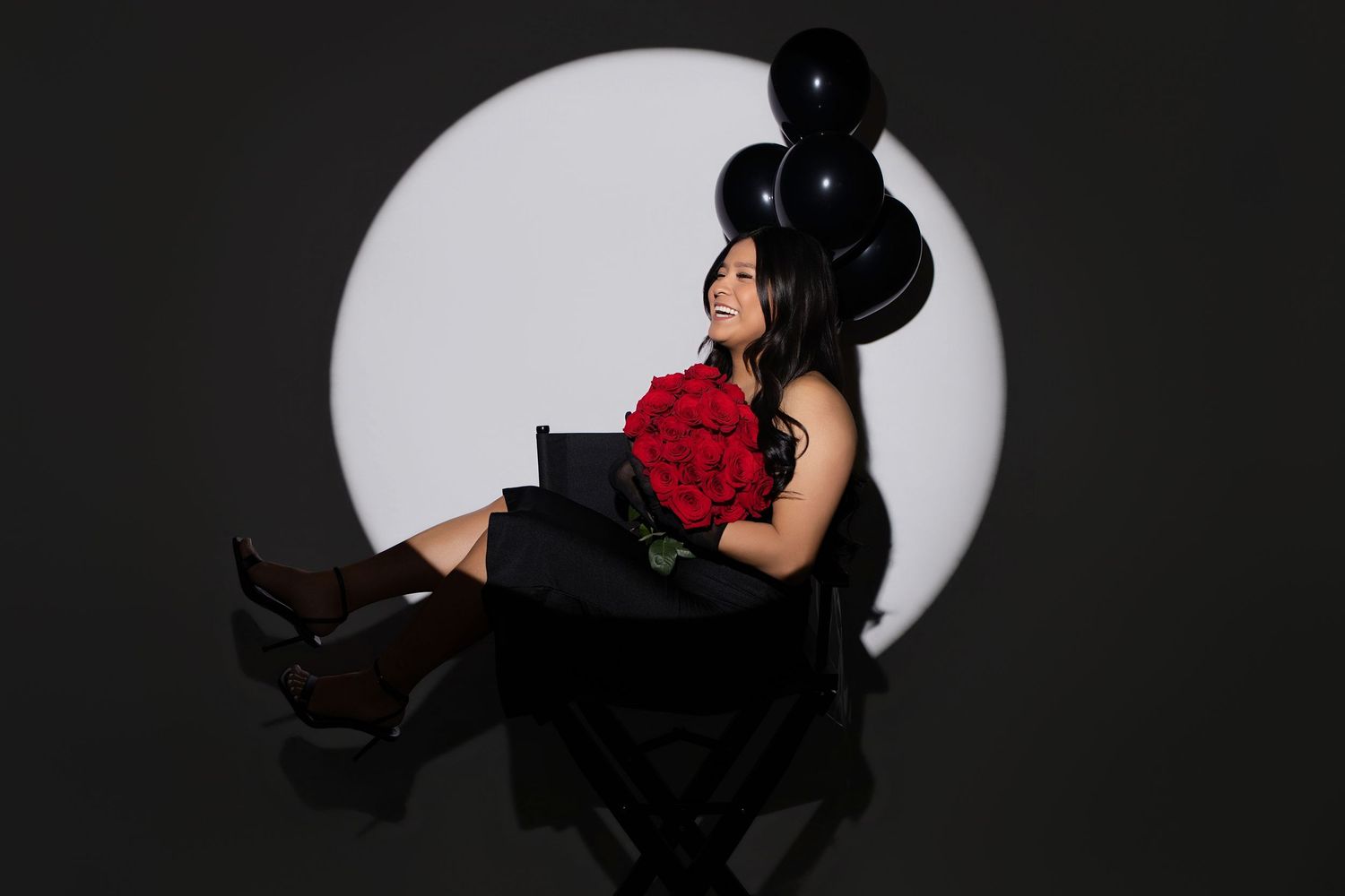 Woman in black dress holding red roses sits in chair with black balloons against white circle backdrop.