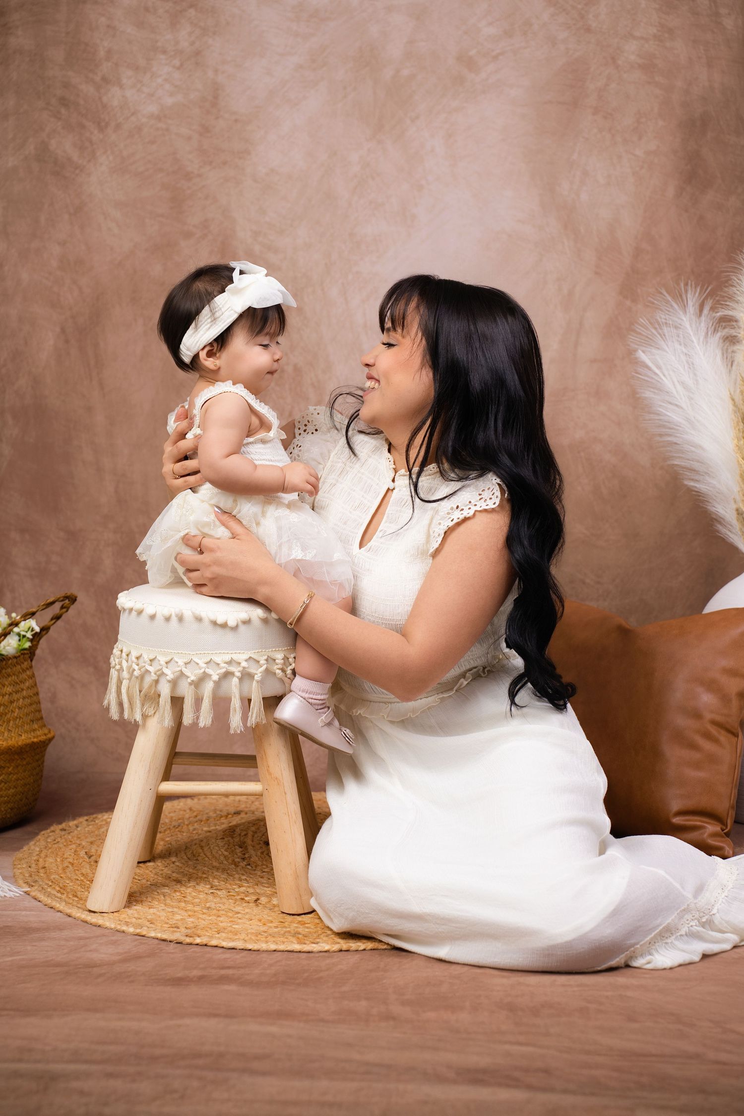 A mother in a white dress smiles at her baby girl in a white outfit, seated on a small stool in a studio.