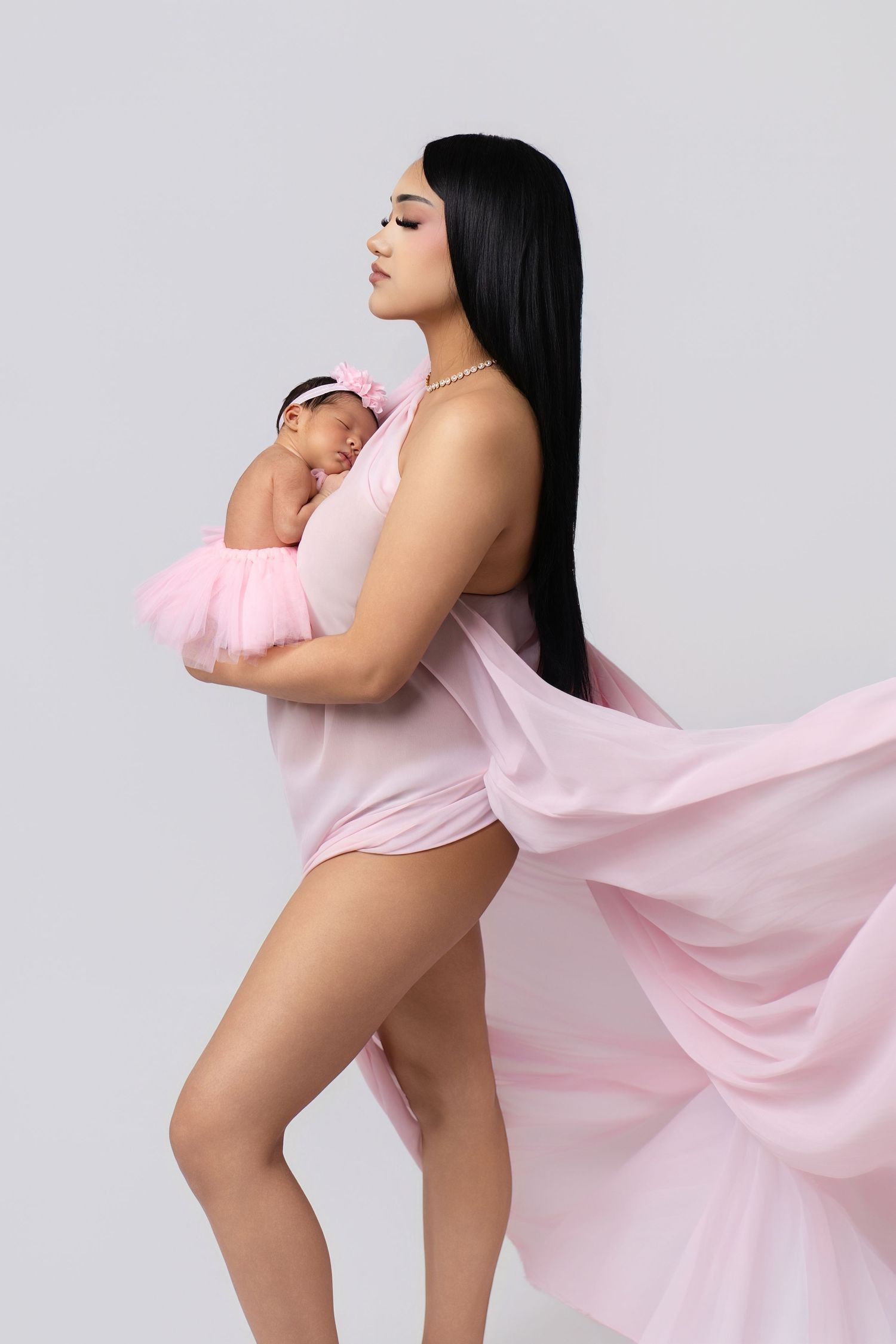 A woman in a flowing pink bodysuit holds her sleeping newborn baby girl dressed in a pink tutu against a white background.