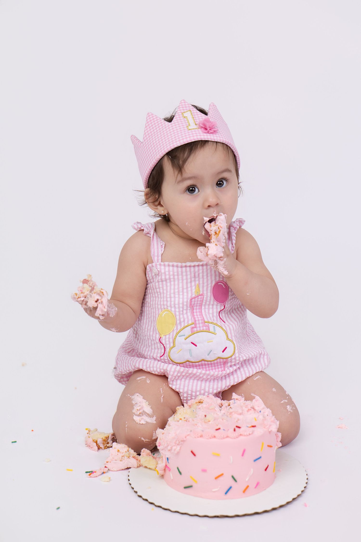 Baby girl in pink crown and outfit smashes first birthday cake, eating frosting with messy hands on white background.
