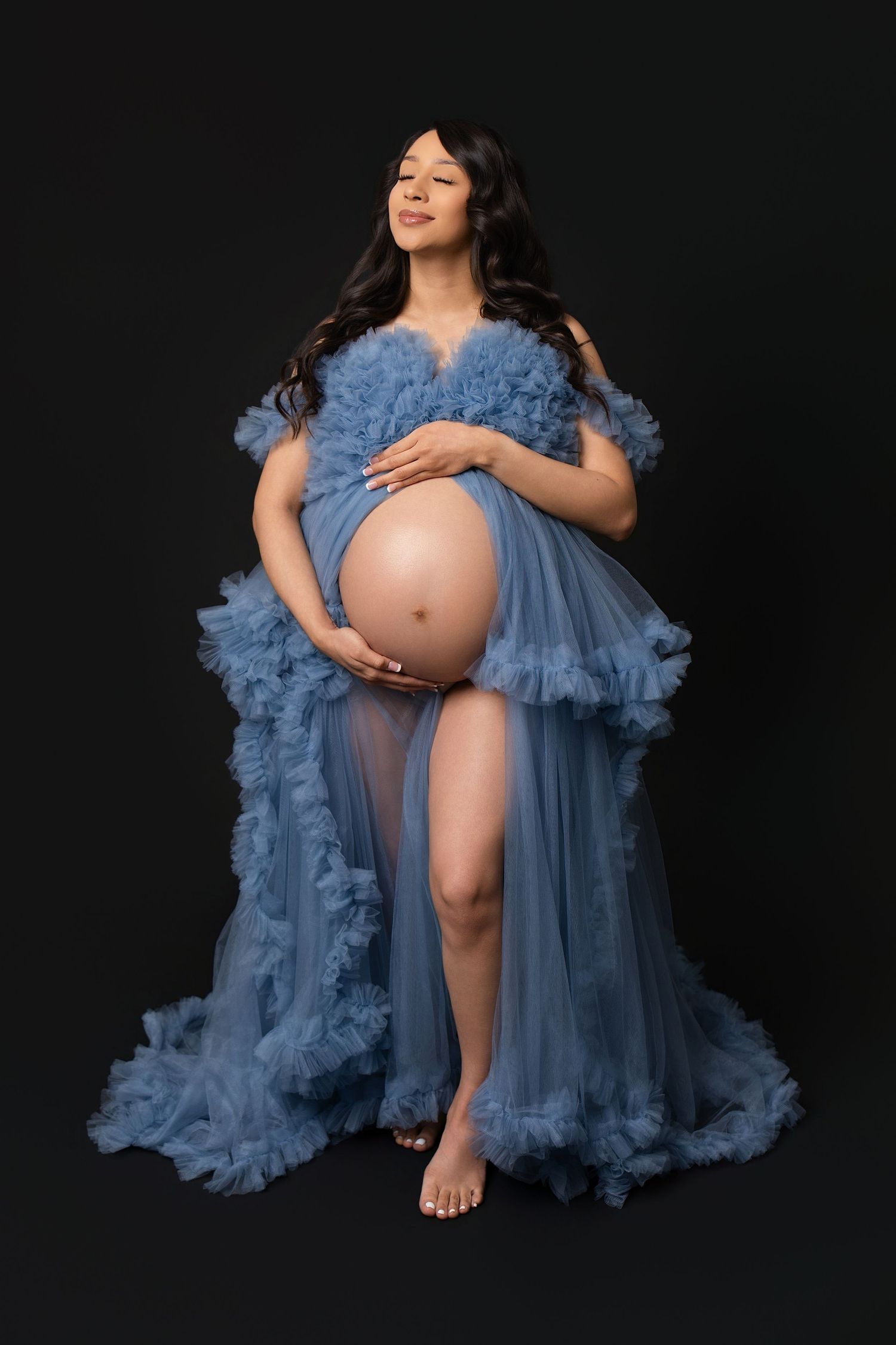 Pregnant woman in blue tulle maternity gown cradling baby bump against black background.