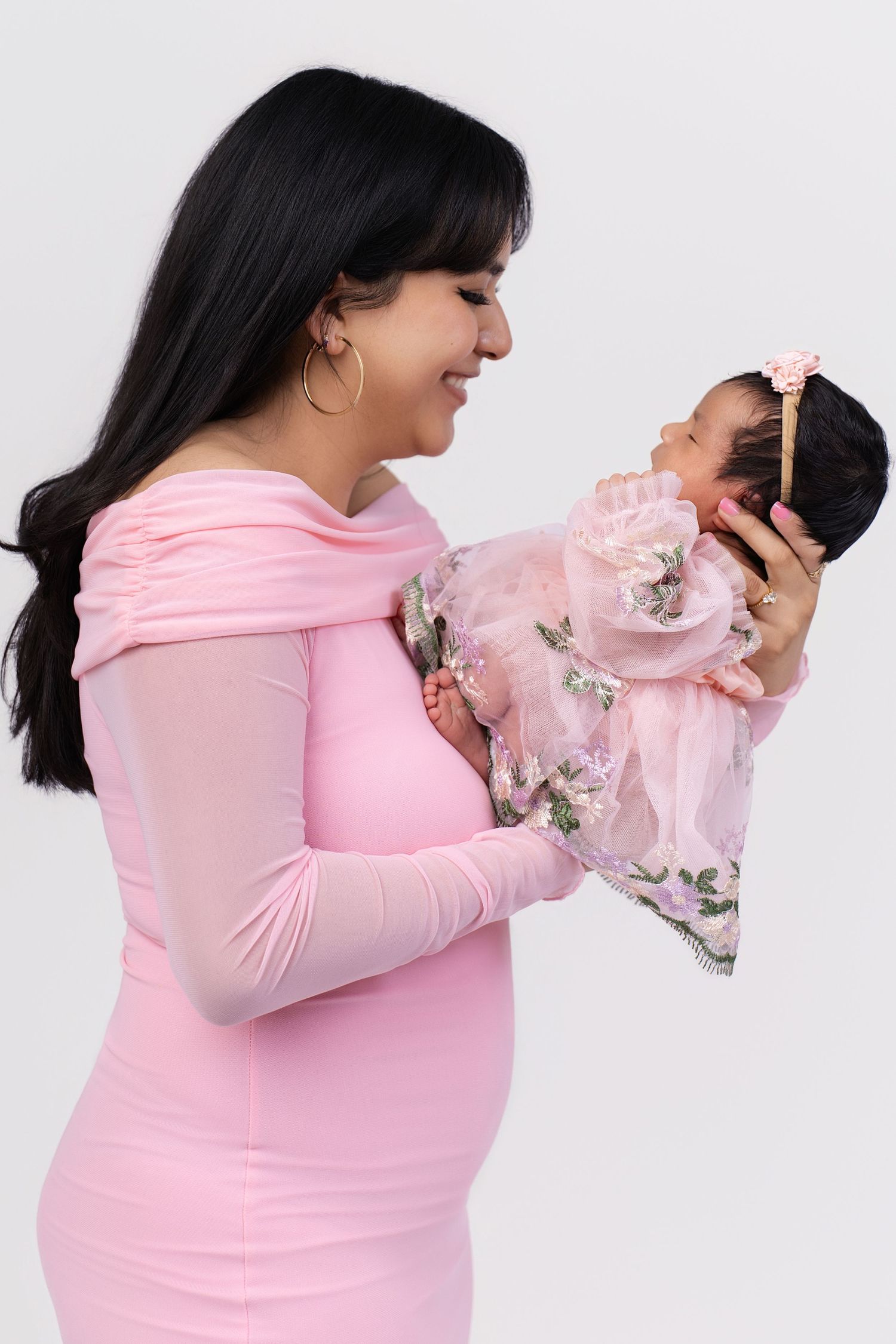 A smiling mother in a pink dress holds her newborn baby dressed in a floral pink outfit against a white background.