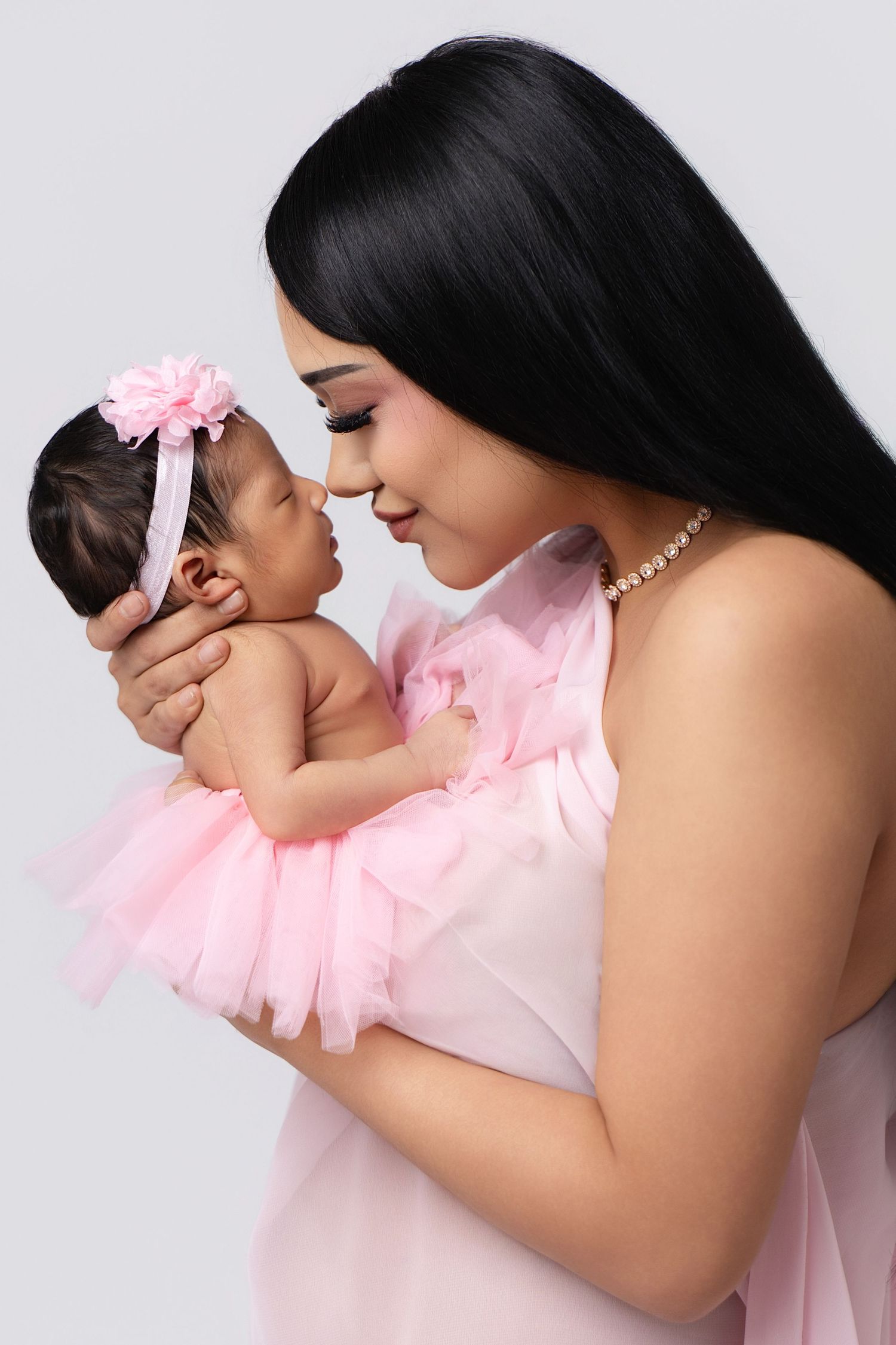 A mother in pink dress tenderly holds her newborn baby girl with a pink floral headband against a white background.