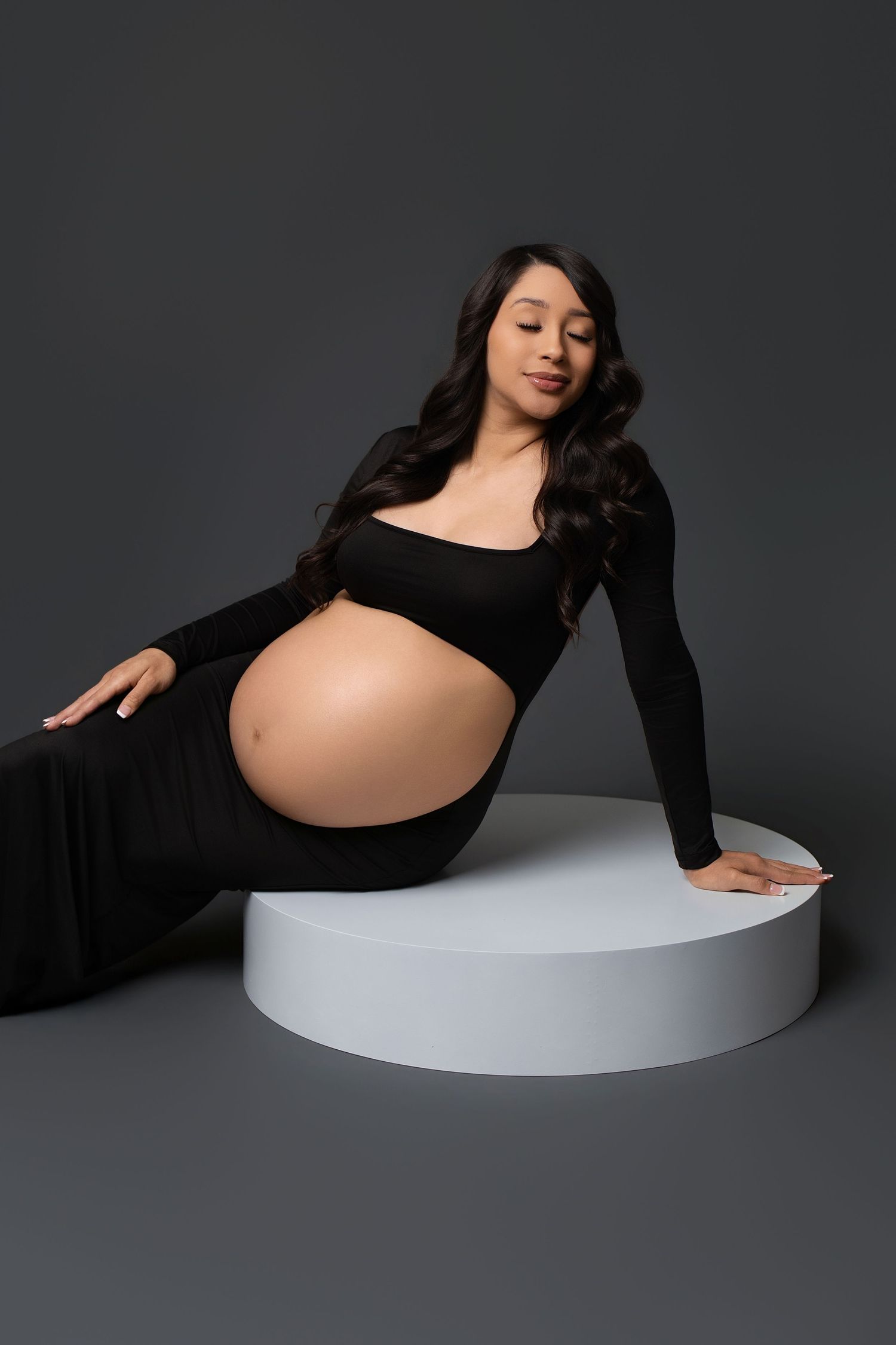 Artistic maternity photo series showing a pregnant figure in black against dark background with white curved platform.