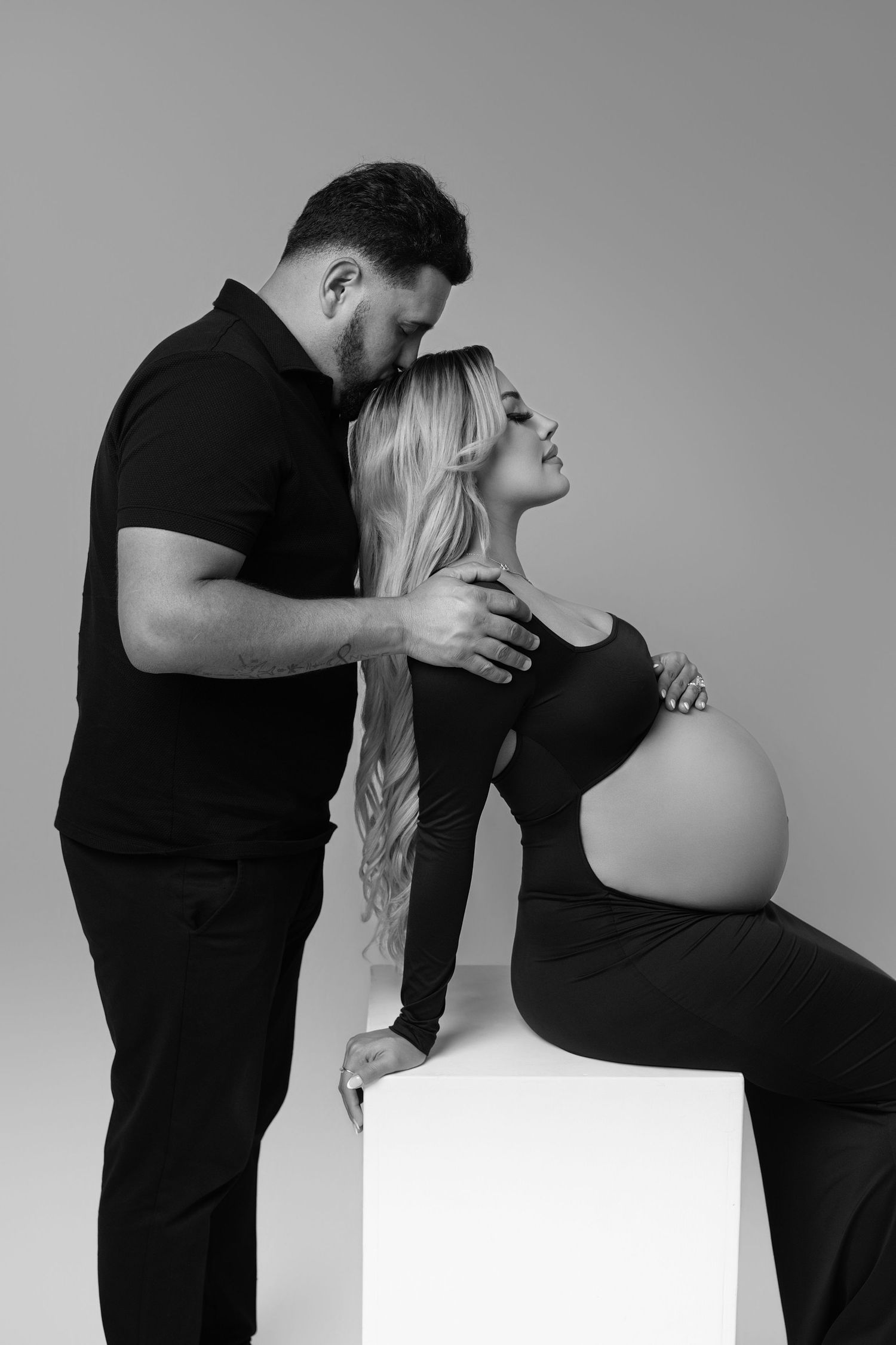 Black and white maternity photo of a couple in black outfits, man kissing pregnant woman's head while she sits on a cube.