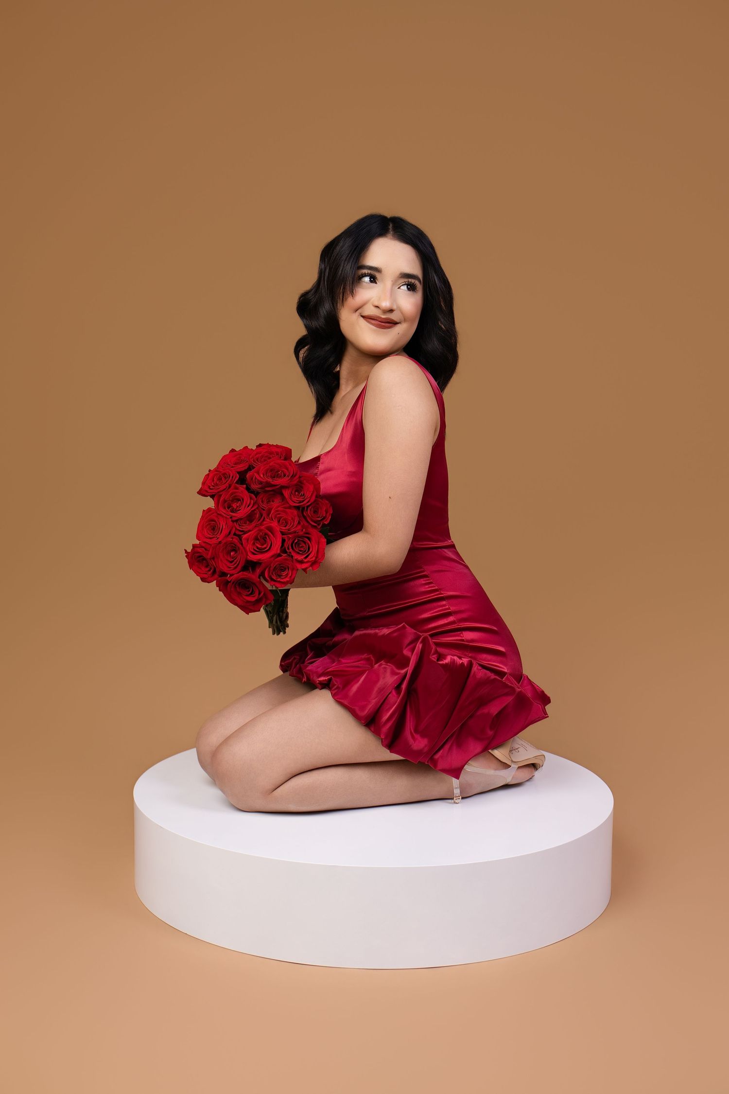 Woman in red satin dress sitting on white platform holding bouquet of red roses on beige background.
