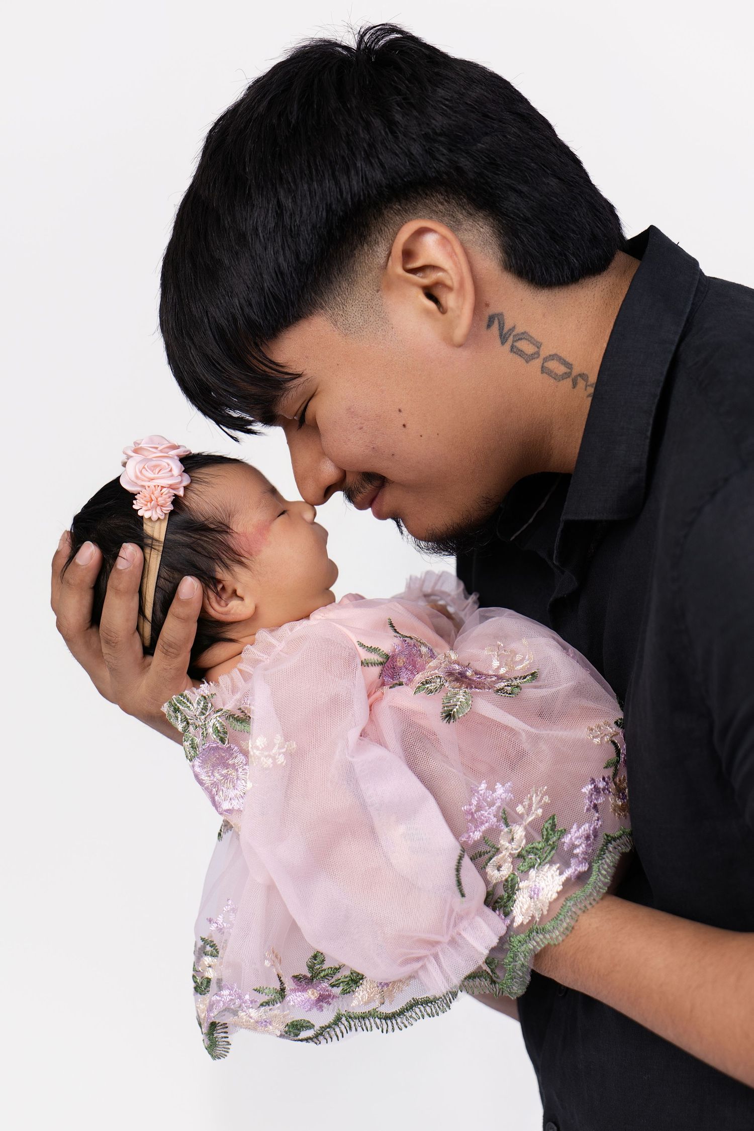 A young father with neck tattoo tenderly kisses his newborn baby girl in a pink floral dress on a white background.