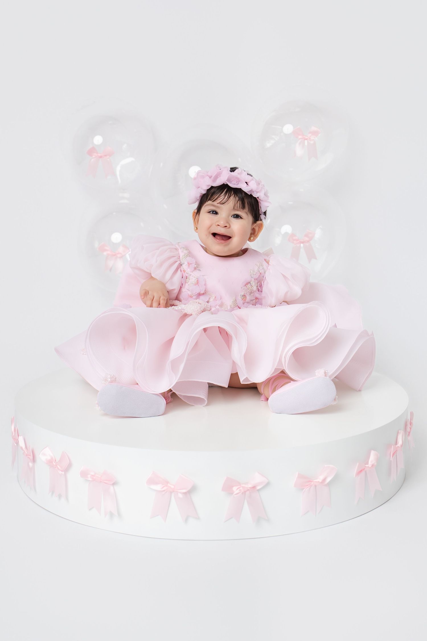 Smiling baby girl in pink ruffled dress sitting on decorated pedestal with pink bow balloons.