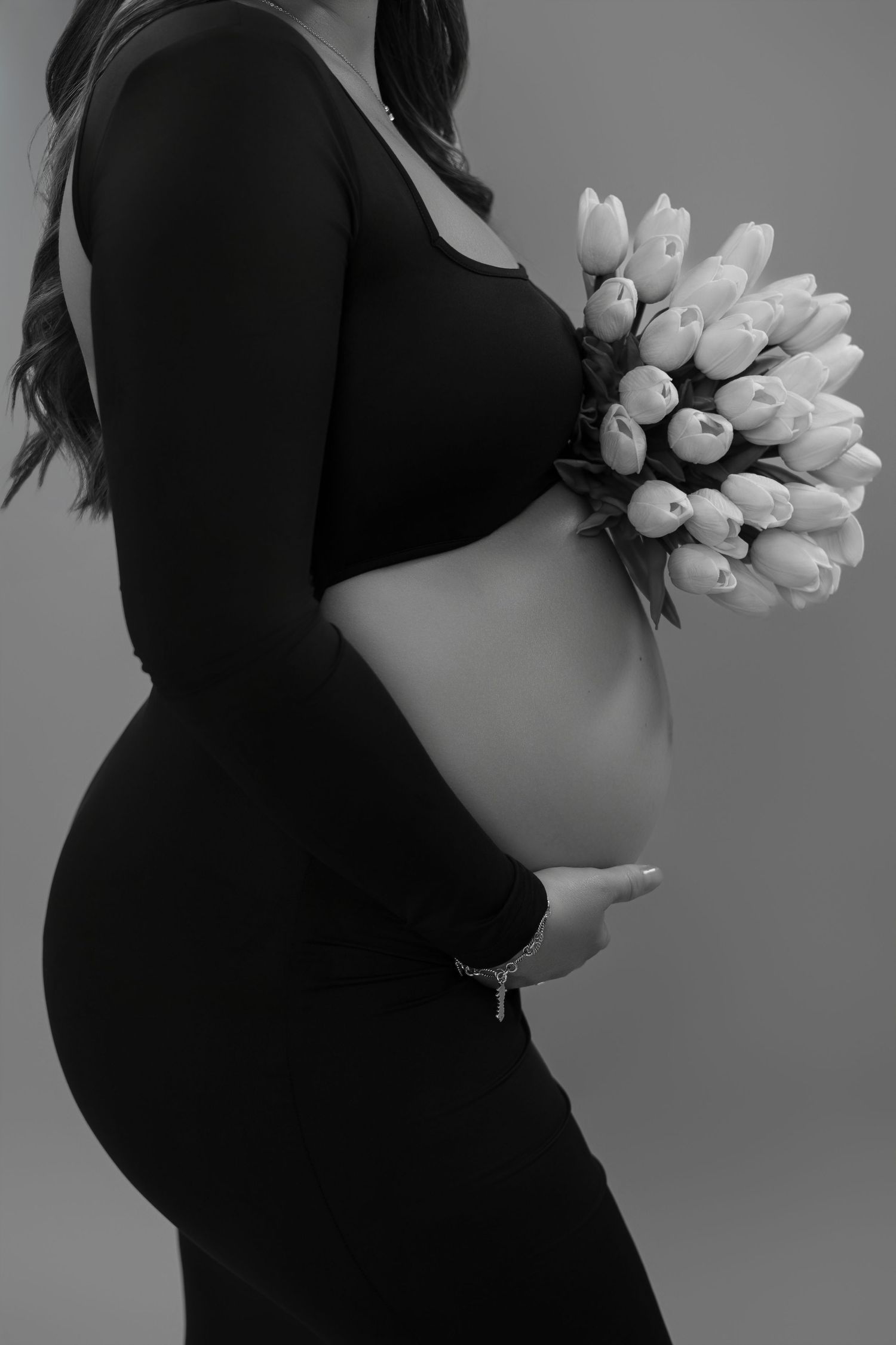 Black and white maternity photo series showing pregnant woman in profile holding white tulips against gray background.