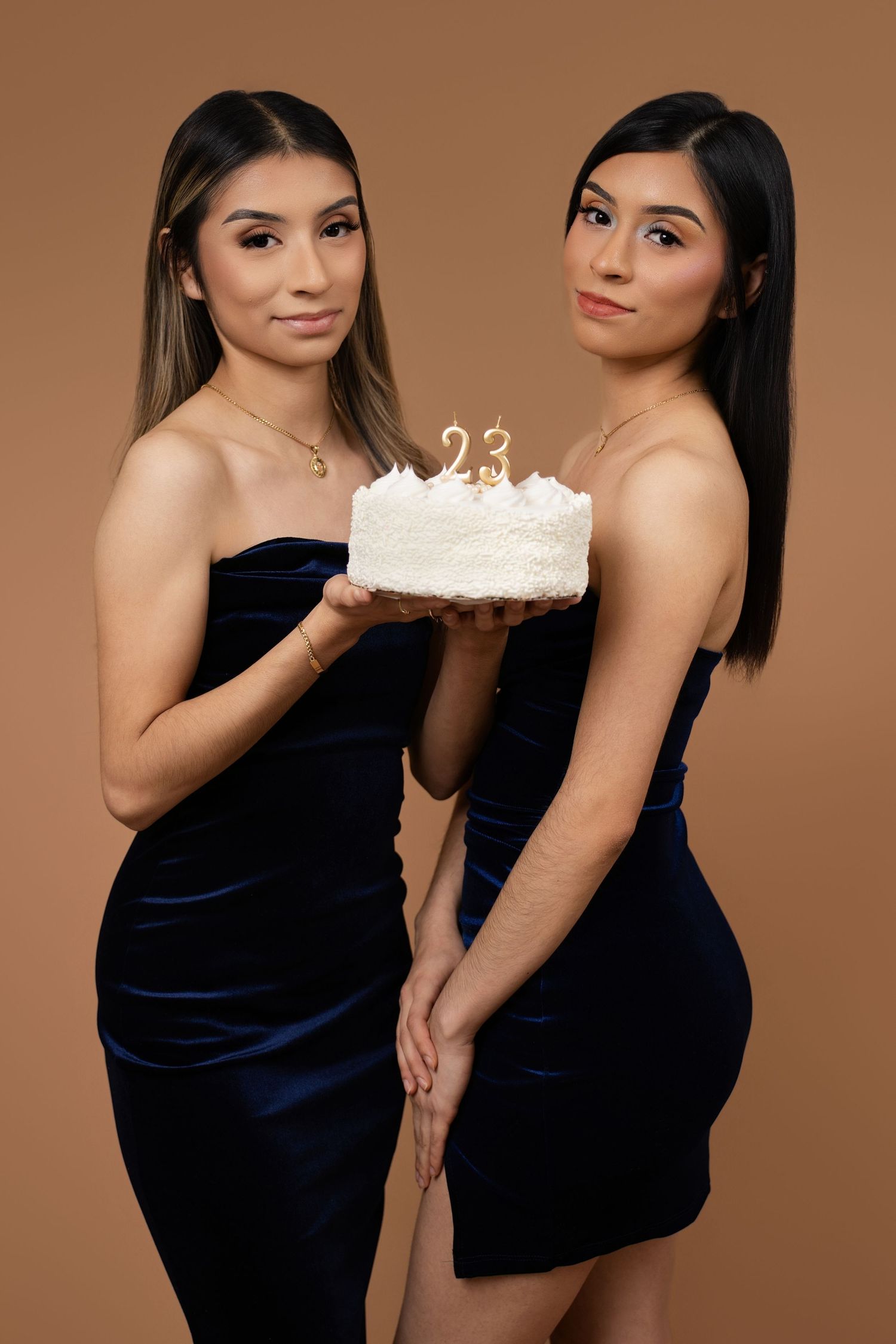 Two young women in navy blue velvet dresses holding a white birthday cake with number 23 candles on beige background.