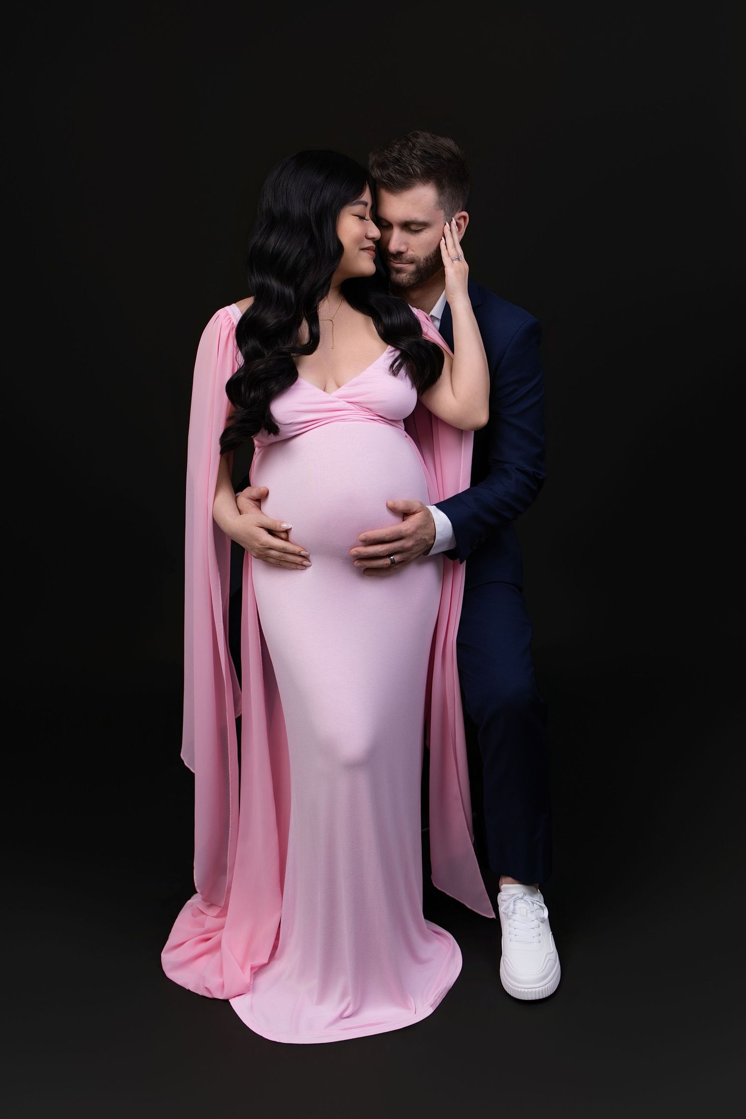 Pregnant woman in pink gown embraced by man in navy suit, romantic maternity photo on black background.