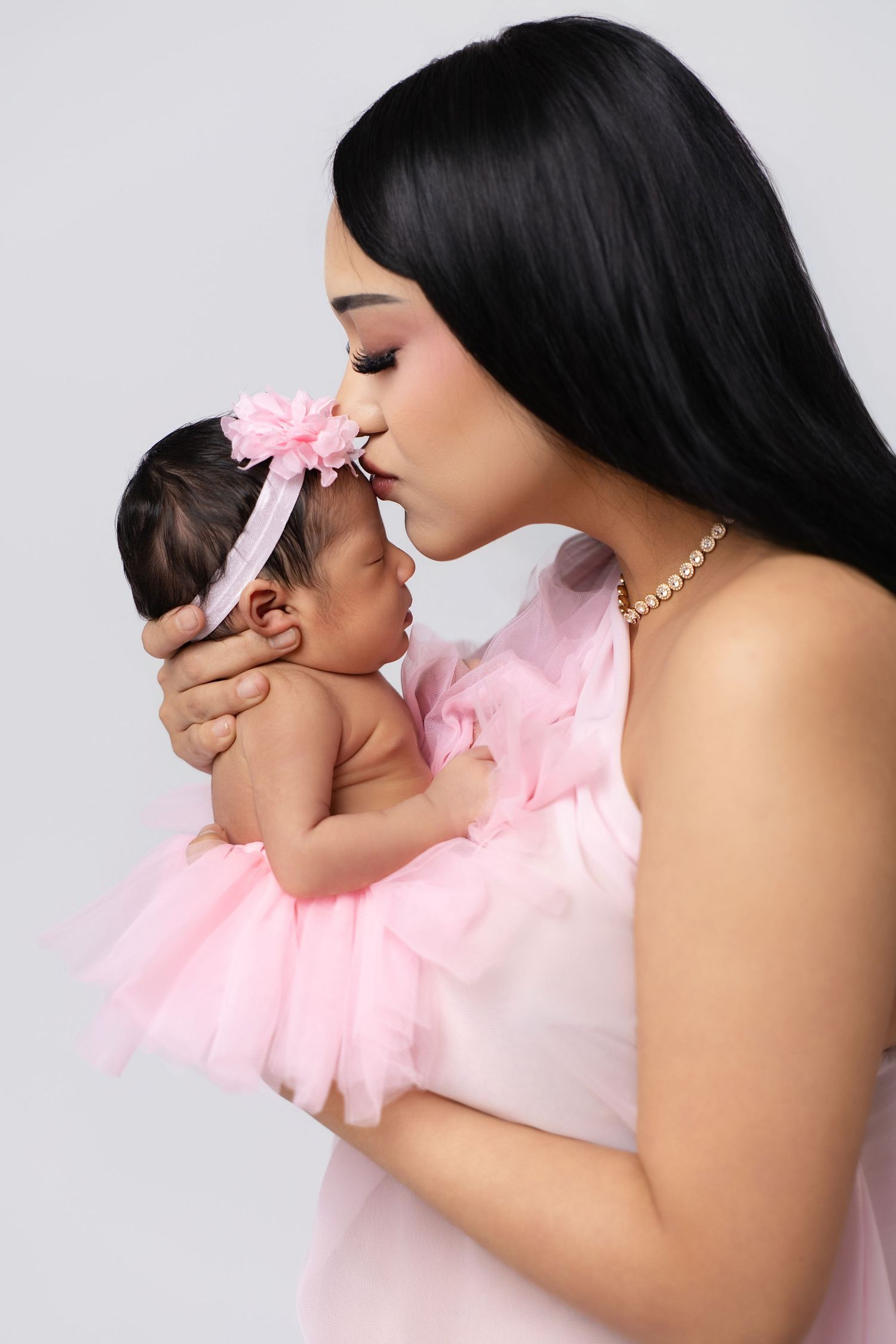 A woman in a pink dress kisses her newborn baby girl wearing a pink tutu and floral headband.
