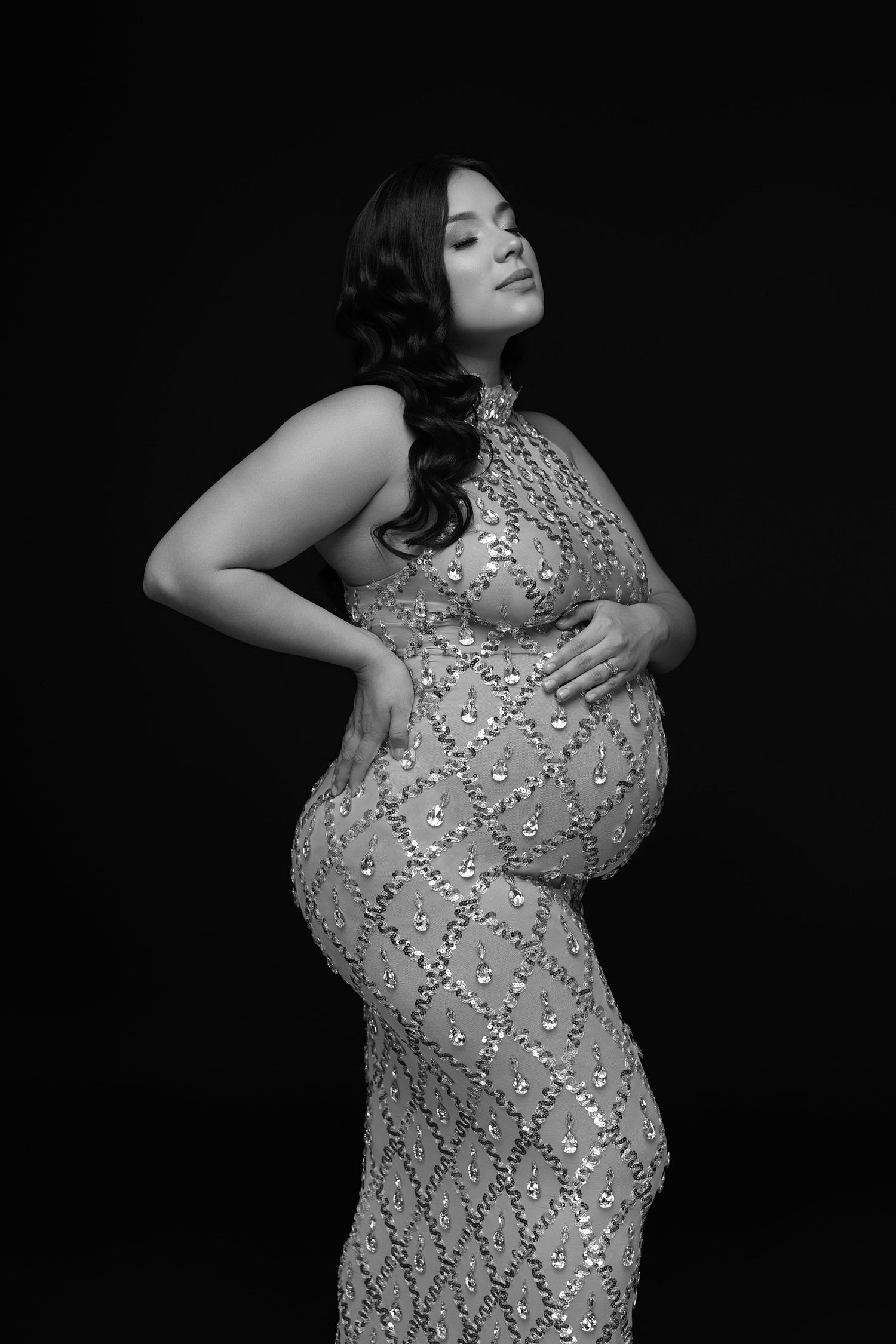 Black and white maternity photo of a pregnant woman in a sequined gown, cradling her belly against a dark background.