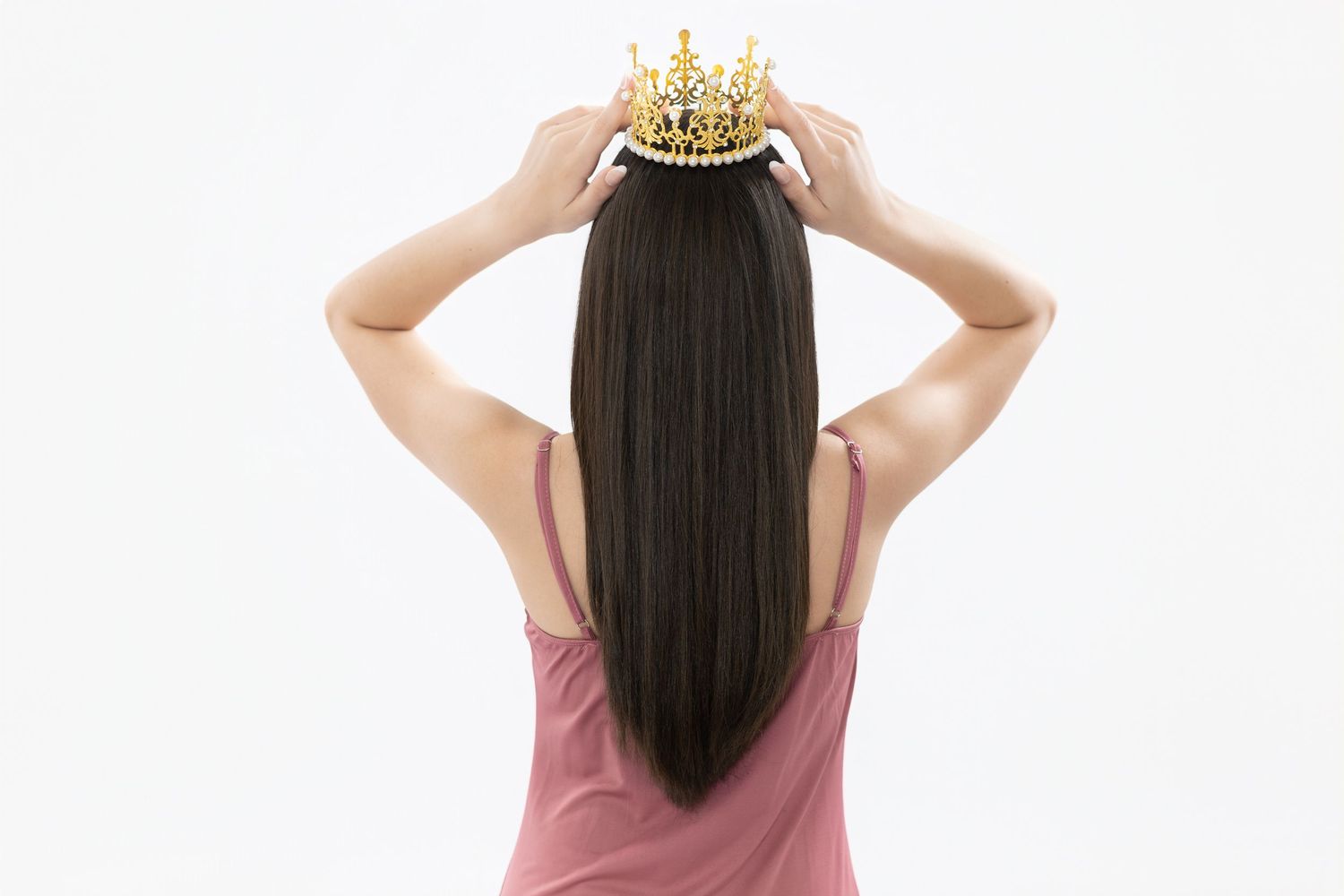 A young woman with long dark hair places a gold crown on her head, viewed from behind on a white background.