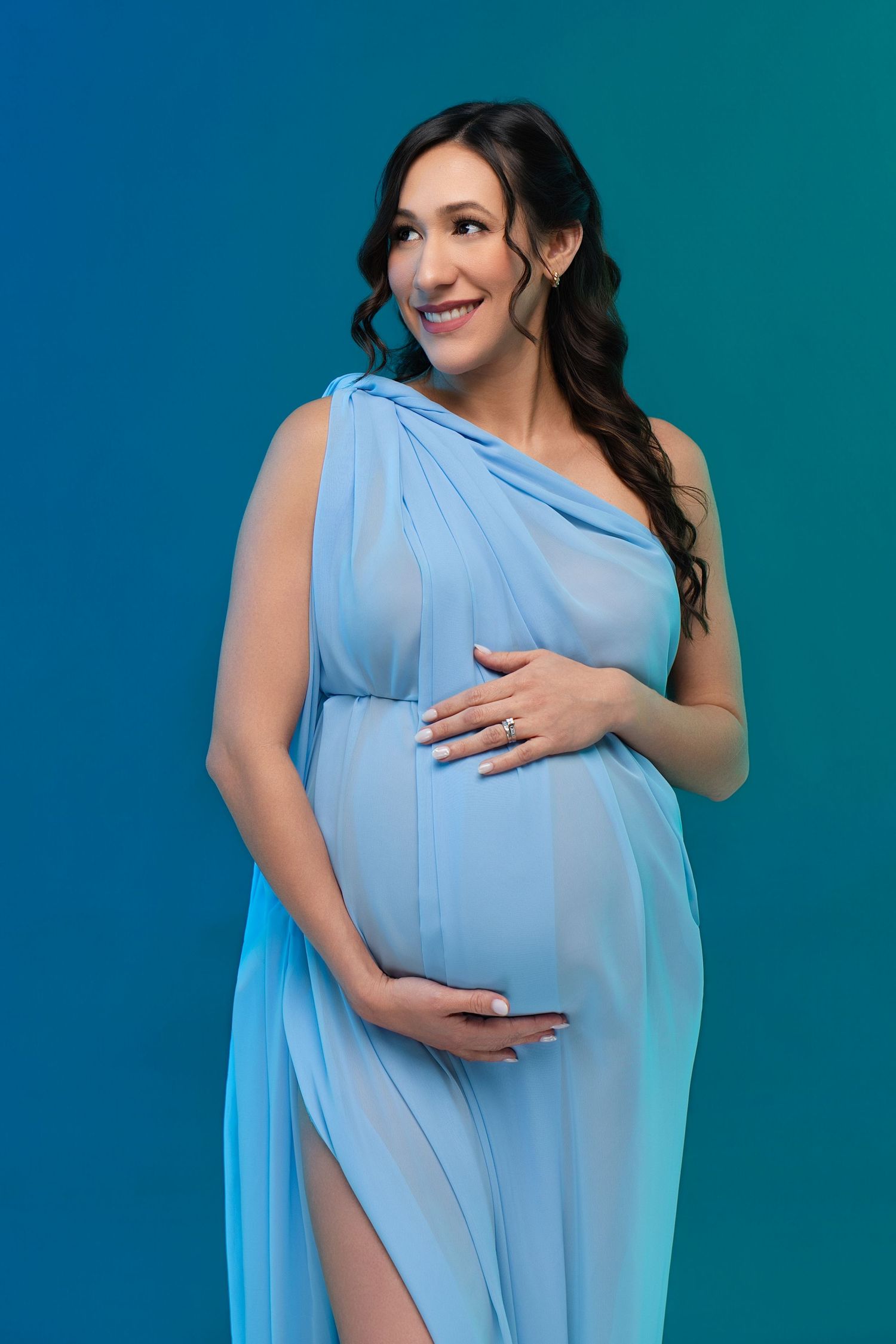 Smiling pregnant woman in elegant blue chiffon dress cradling her baby bump against a blue gradient background.