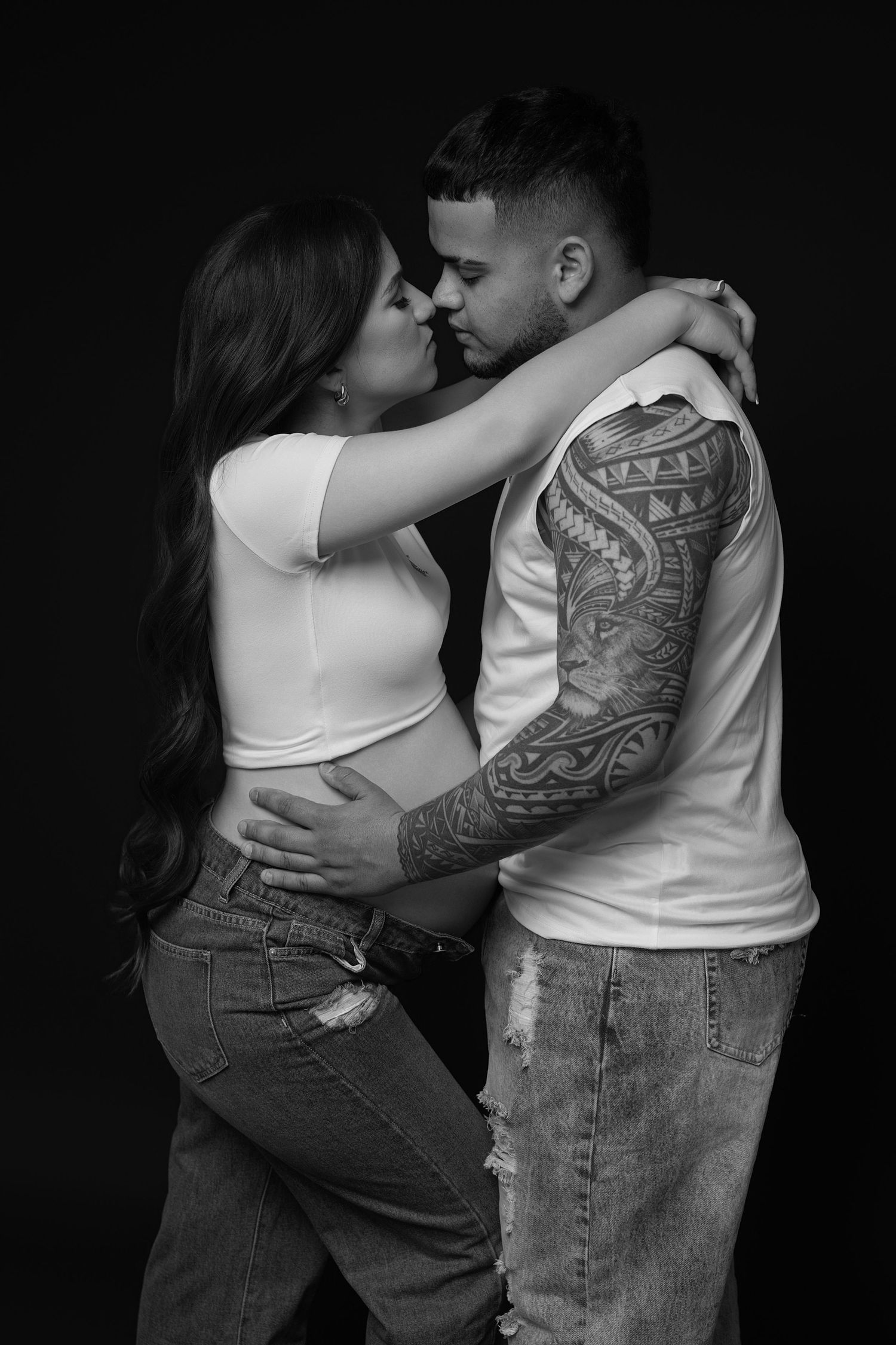 Black and white photo of a tattooed couple embracing intimately, the woman visibly pregnant, against a dark studio background.