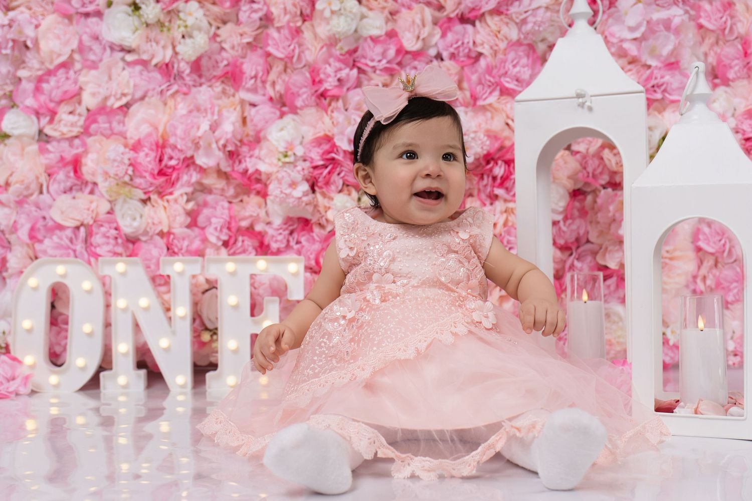 Baby girl in pink lace dress sits before floral wall with ONE marquee letters celebrating her first birthday.
