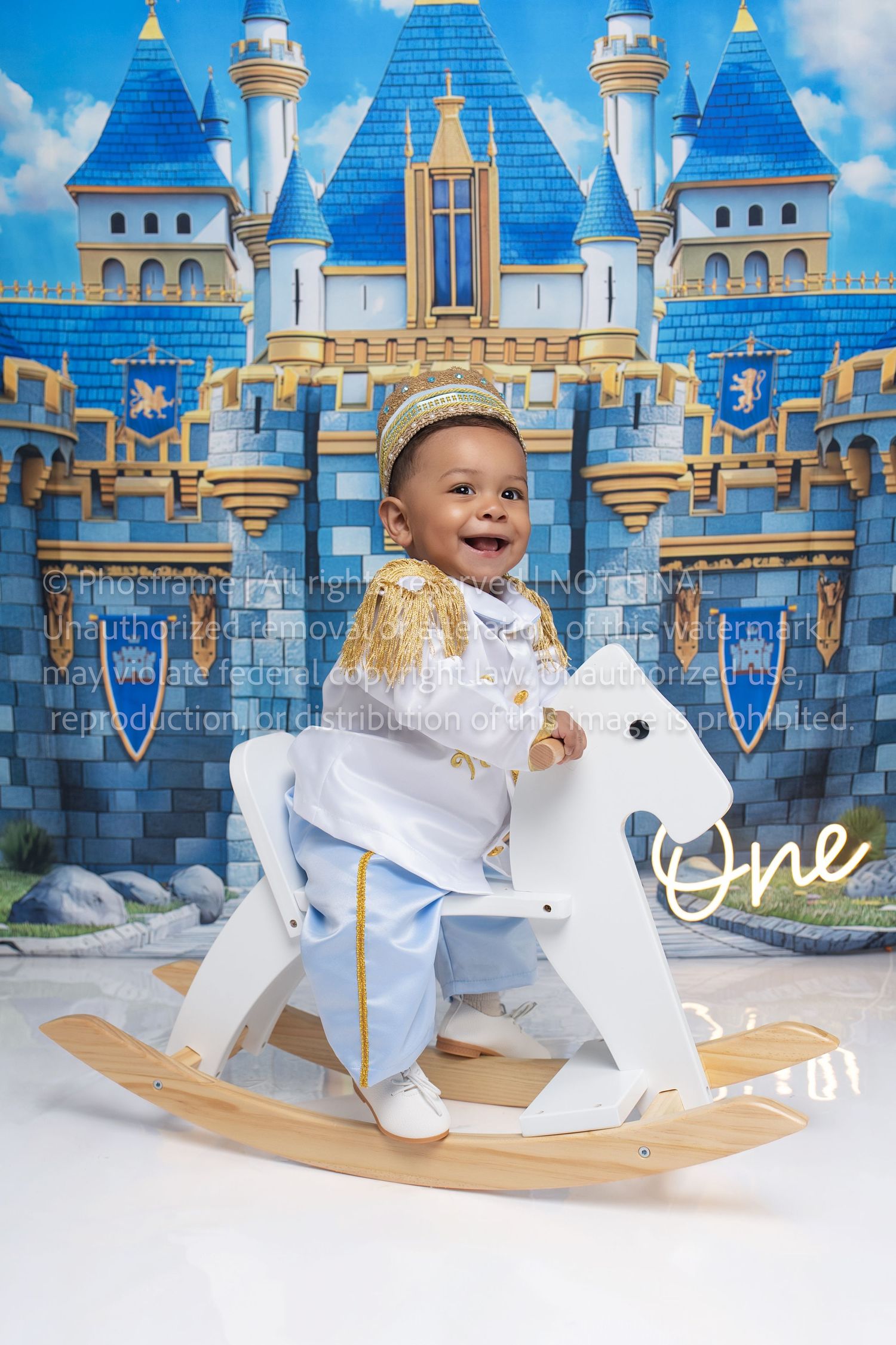 Smiling baby boy in prince outfit sits on white rocking horse with blue castle backdrop celebrating first birthday.